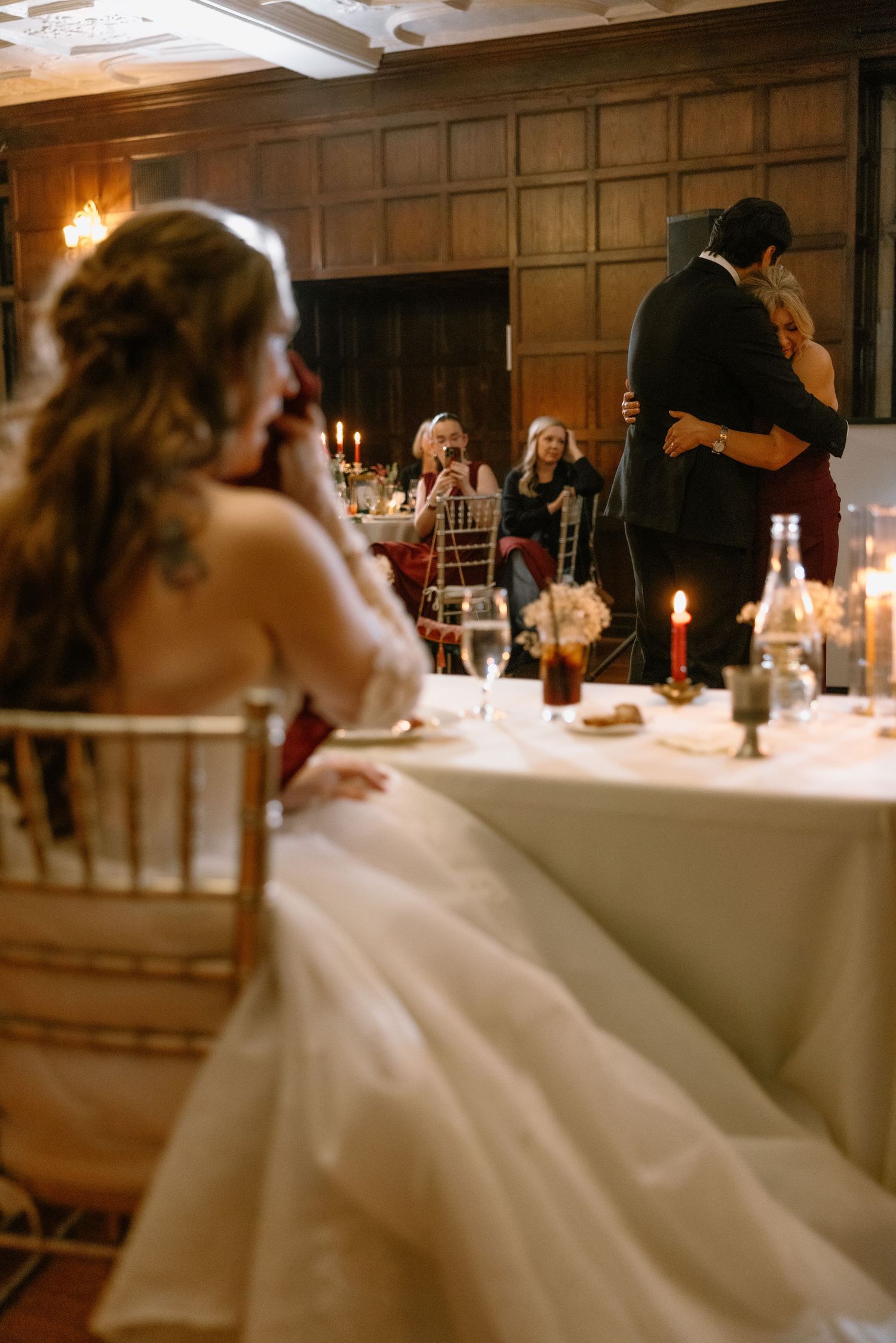 Bride in a flowing ivory gown watches from her seat as guests embrace and dance beside a candlelit reception table in a wood-paneled venue.