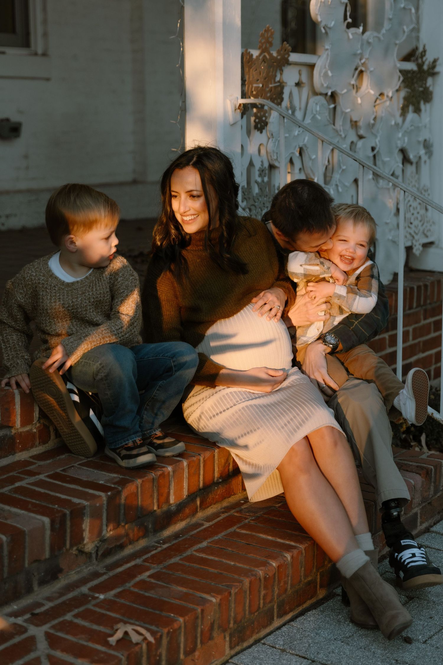 A happy family with two toddlers sits on brick porch steps in warm golden light, mom showing a baby bump.