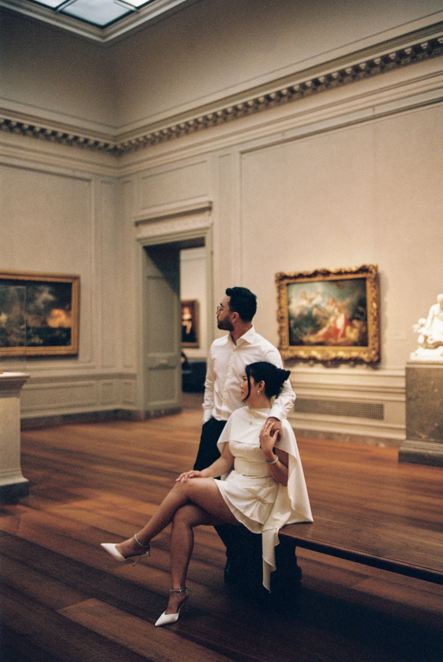 Couple posing elegantly in a classic art museum gallery with ornate paintings and hardwood floors.