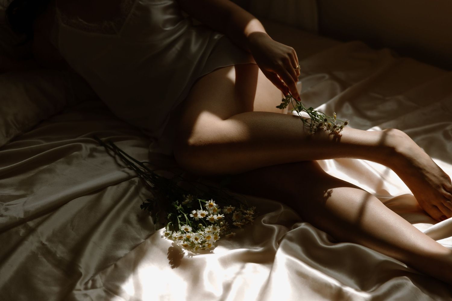 A person in a short dress sits on silky bedding, holding delicate white wildflowers in soft, moody natural light.