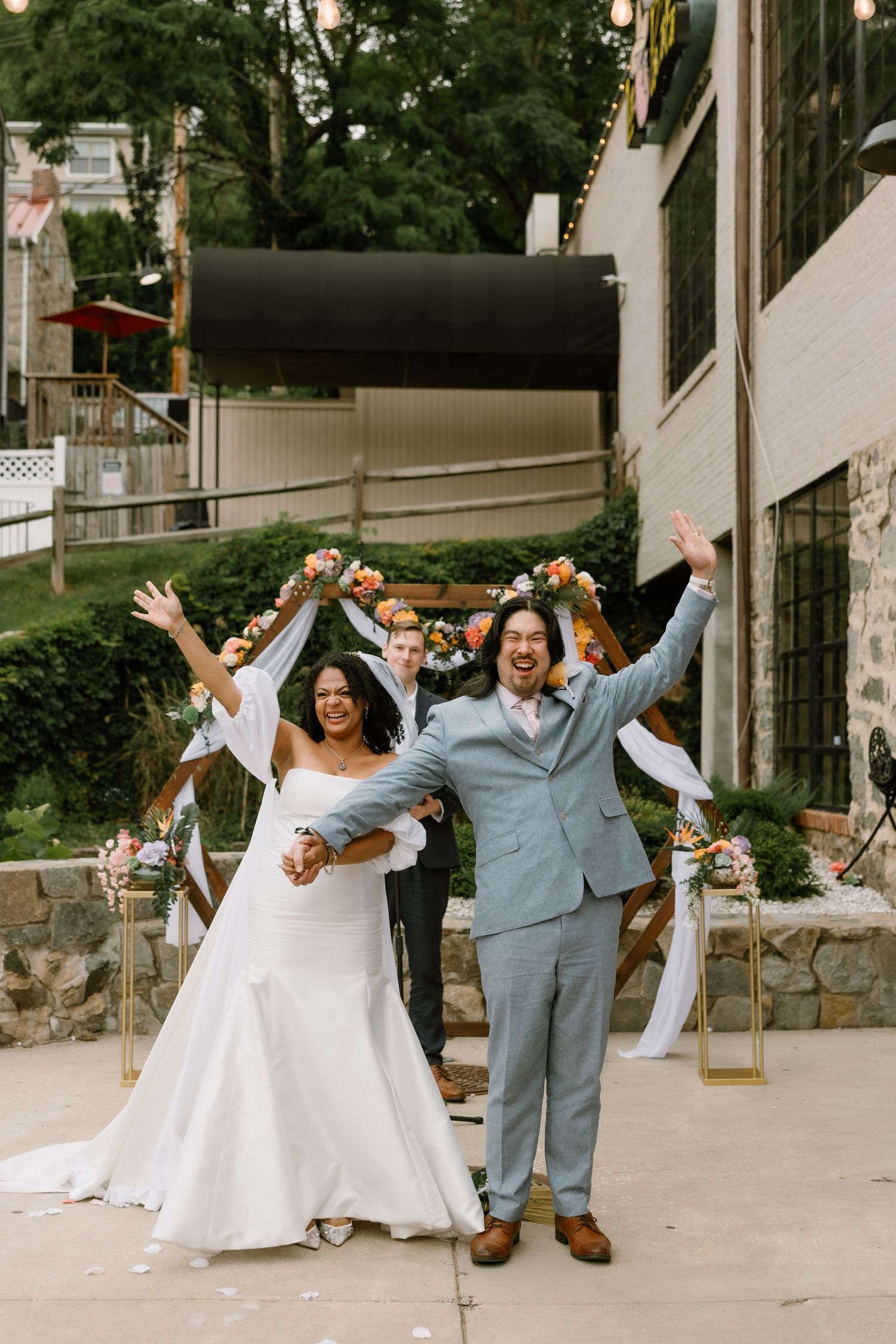 Joyful newlyweds celebrate after outdoor wedding ceremony, bride in white gown and groom in light blue suit, arms raised.