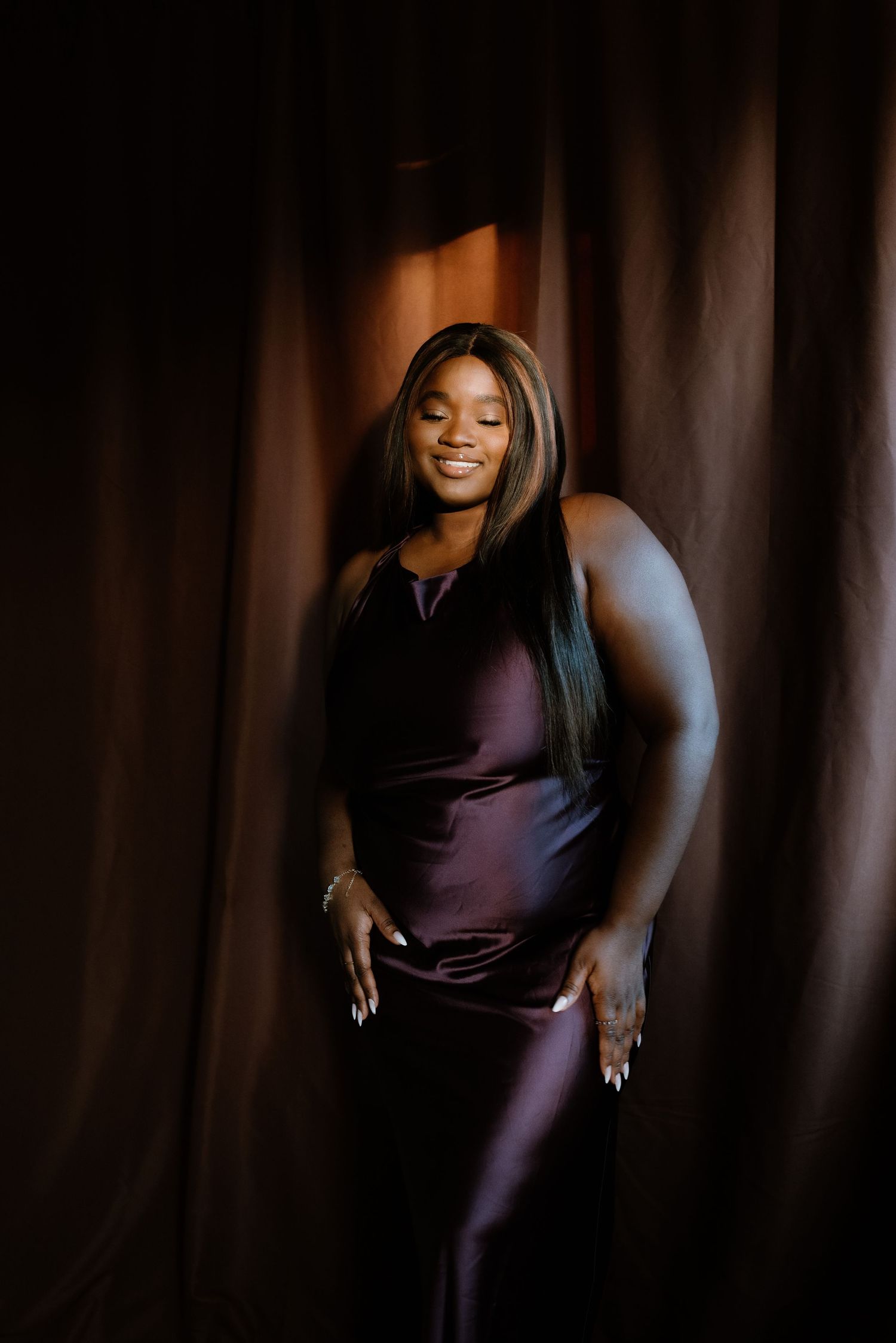 A woman in a purple satin gown poses gracefully against brown drapes in warm studio lighting.