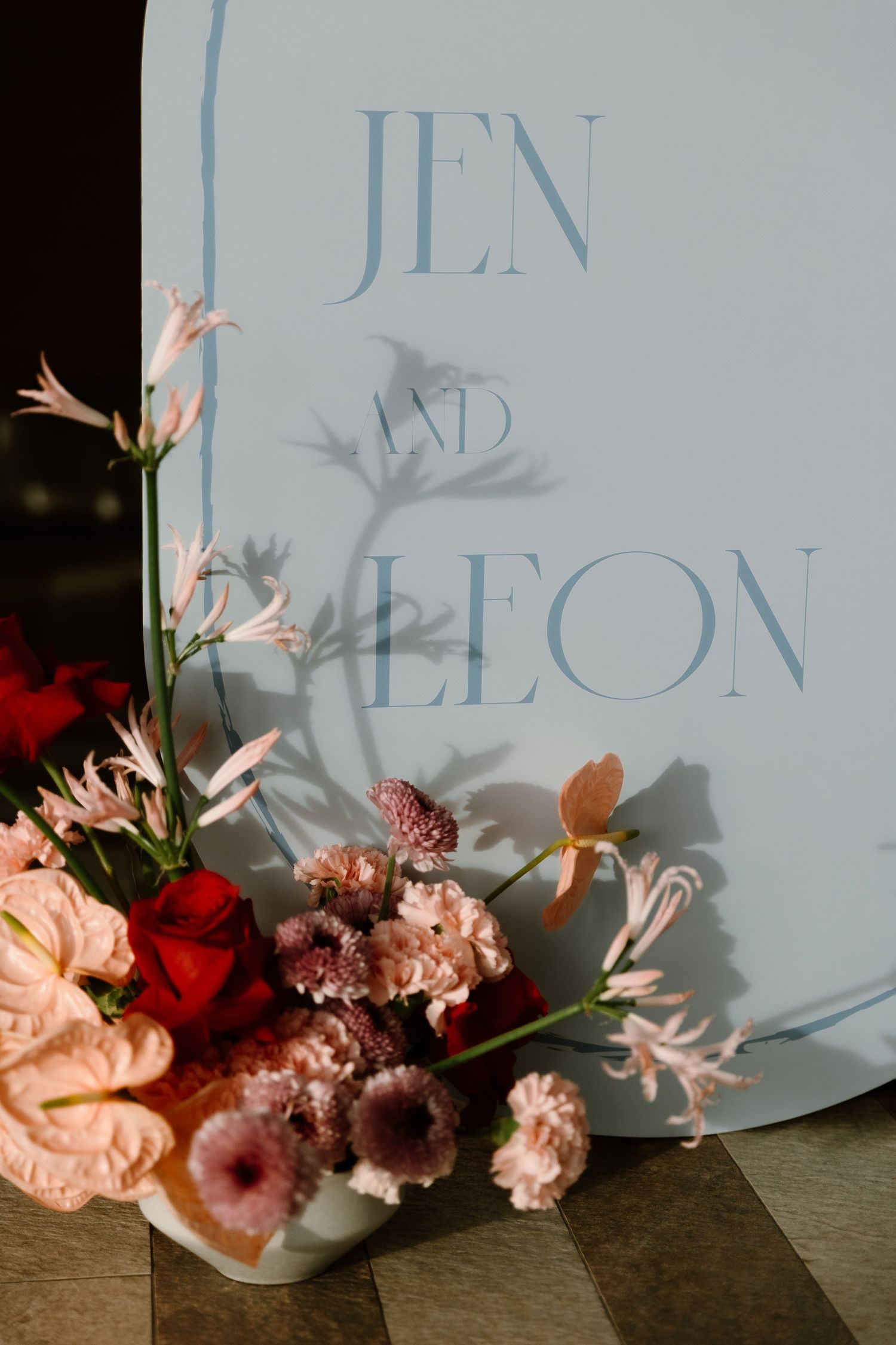 Elegant wedding sign reading Jen and Leon adorned with pink, red, and blush floral arrangements on a light blue backdrop.