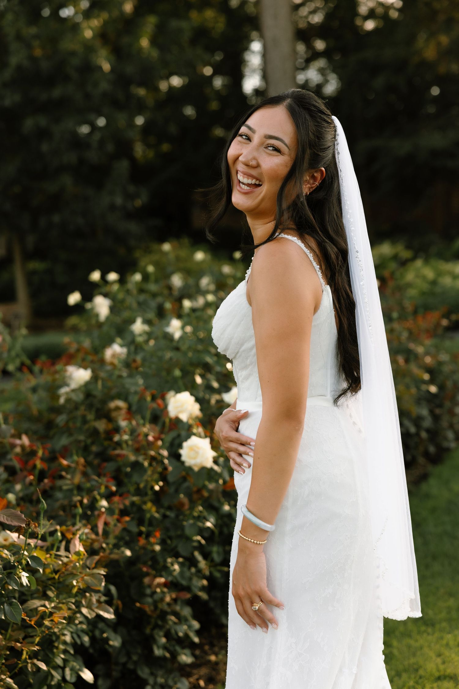 Smiling bride in white wedding dress and veil poses in garden with blooming roses.