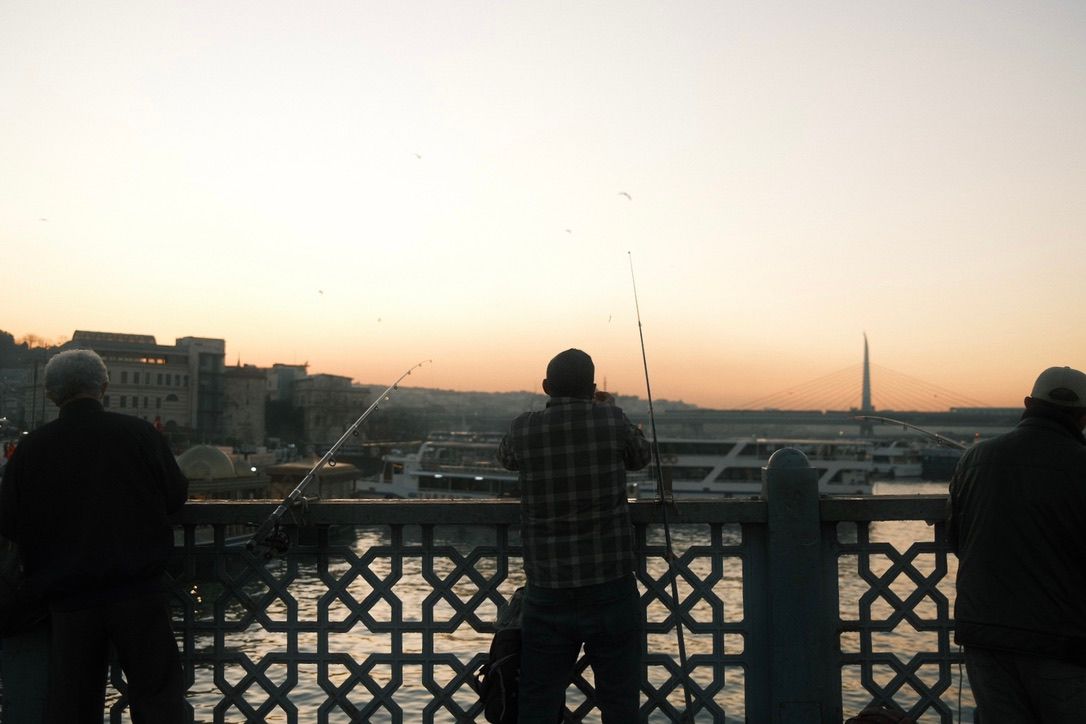 Three silhouetted fishermen casting lines from a bridge railing at sunset over a calm waterway with city skyline.