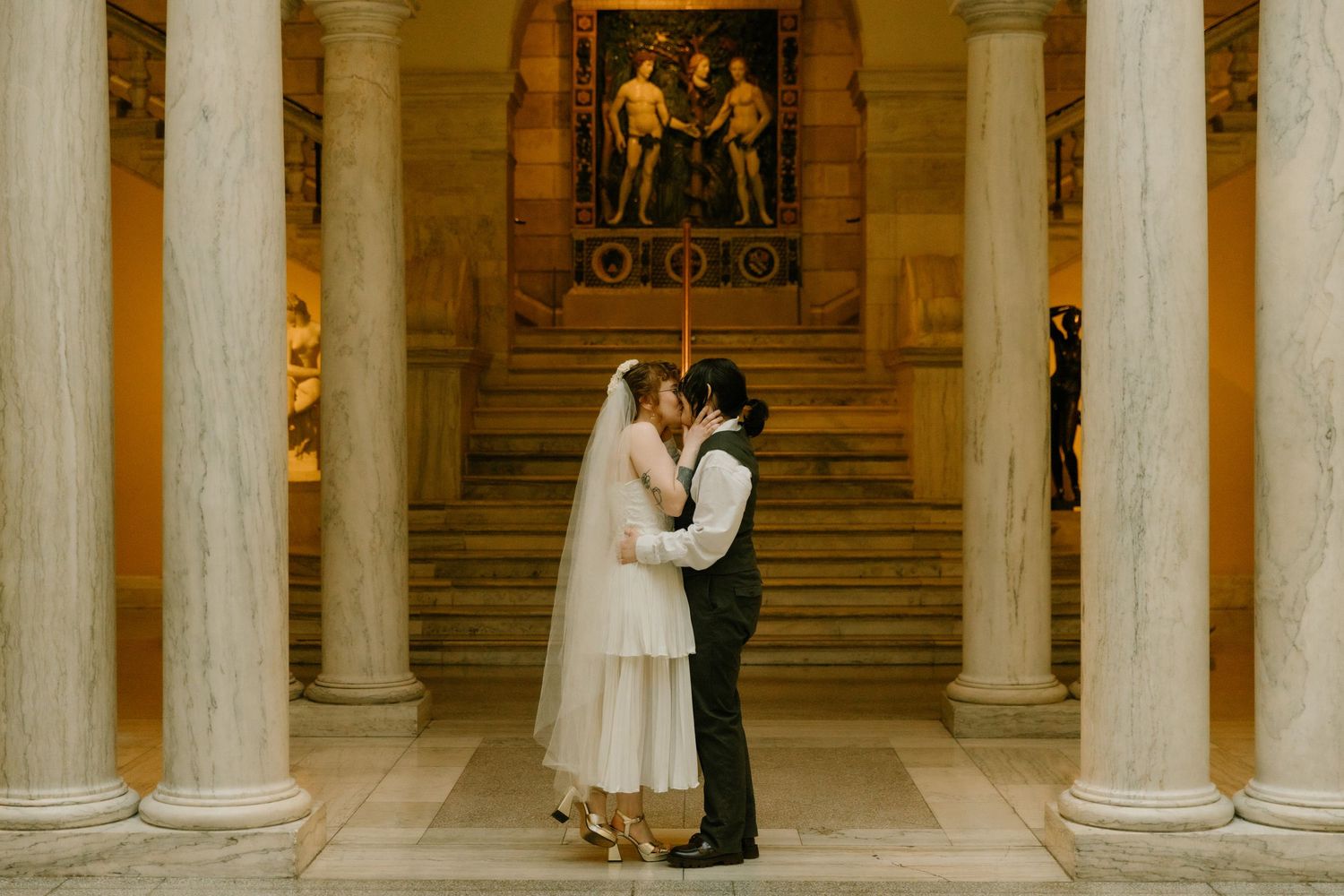 Bride and groom share a romantic kiss inside a grand hall with tall columns and classical mural backdrop.