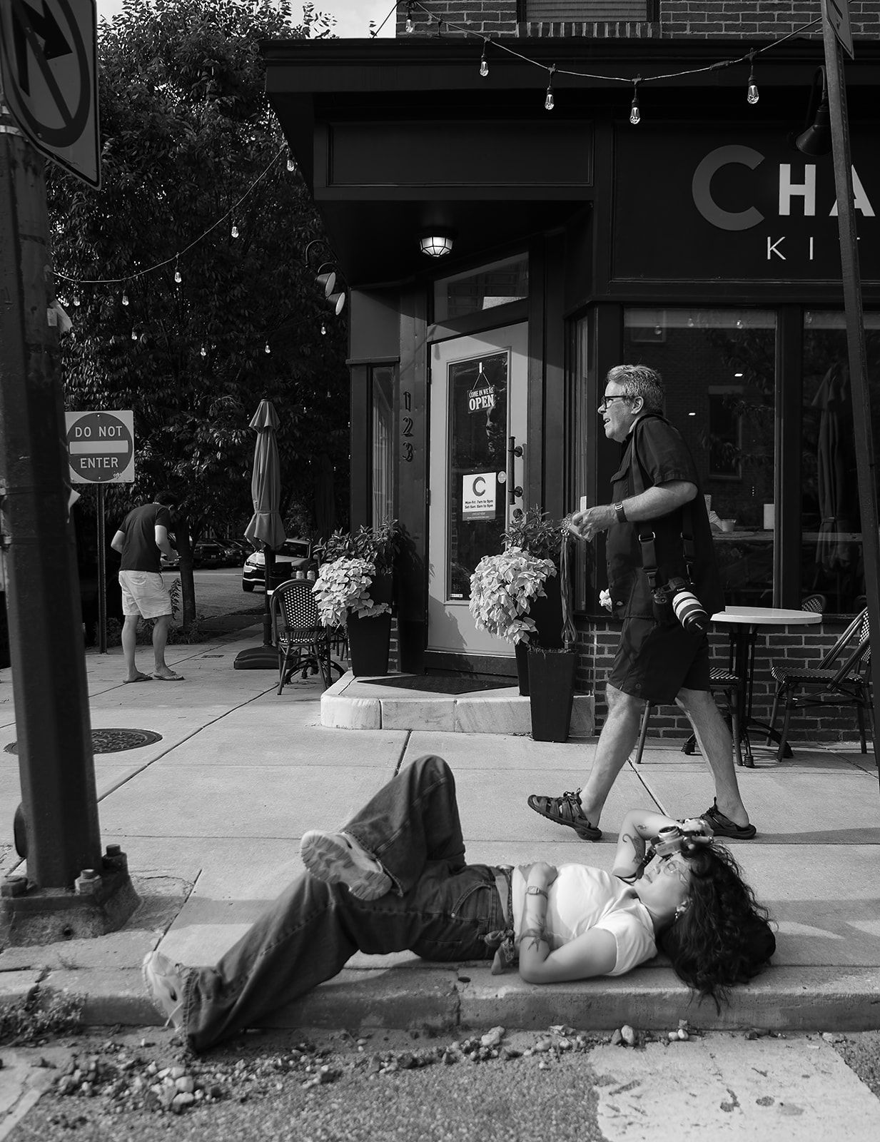 Black and white street photo of a person lying on a sidewalk while a man walks past a storefront.