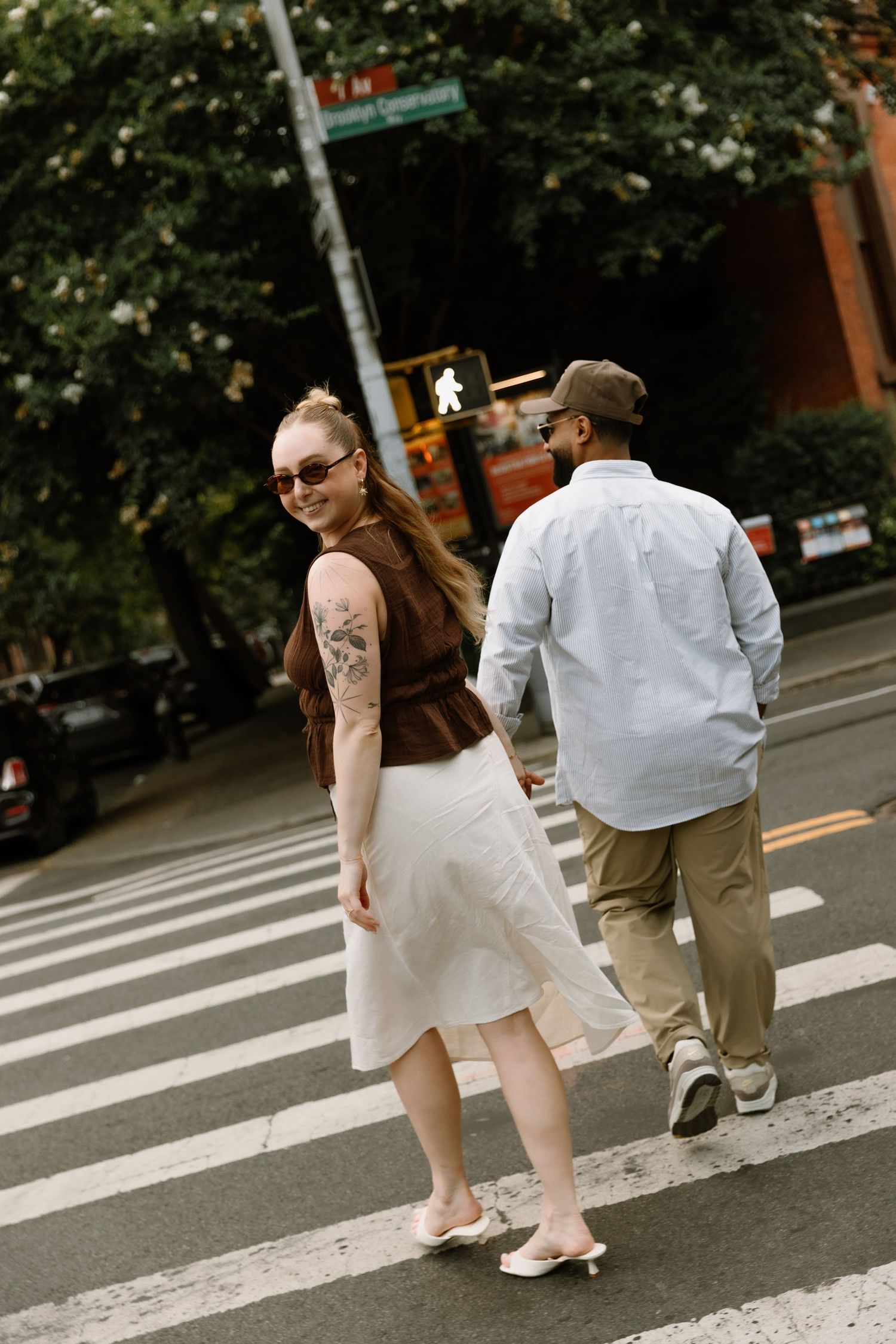 A couple crosses a city street crosswalk, woman in white skirt and brown top with tattoo, man in light blue shirt.