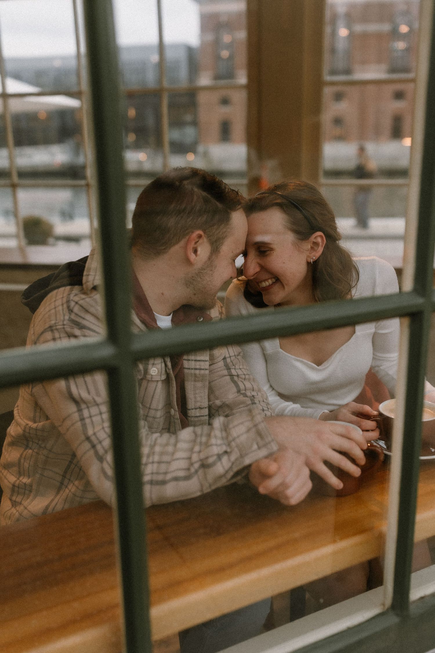 A couple shares a romantic moment at a cafe table, photographed through a window with city waterfront views.