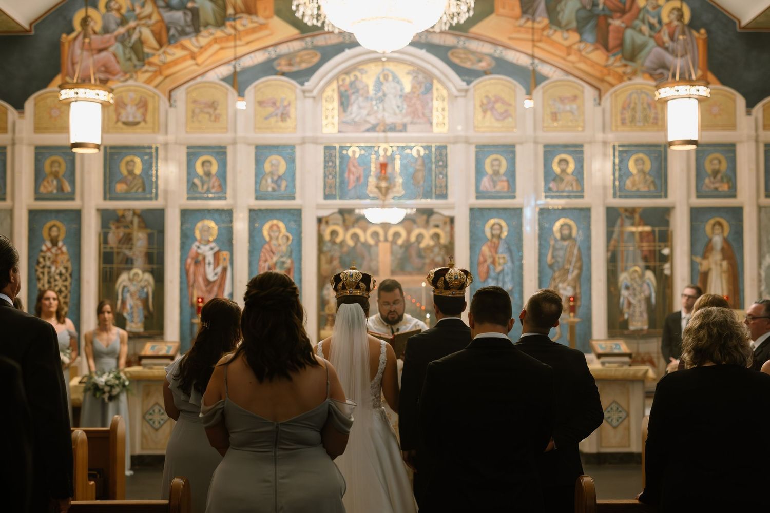 Orthodox church wedding ceremony with bride and groom at ornate iconostasis altar surrounded by guests.
