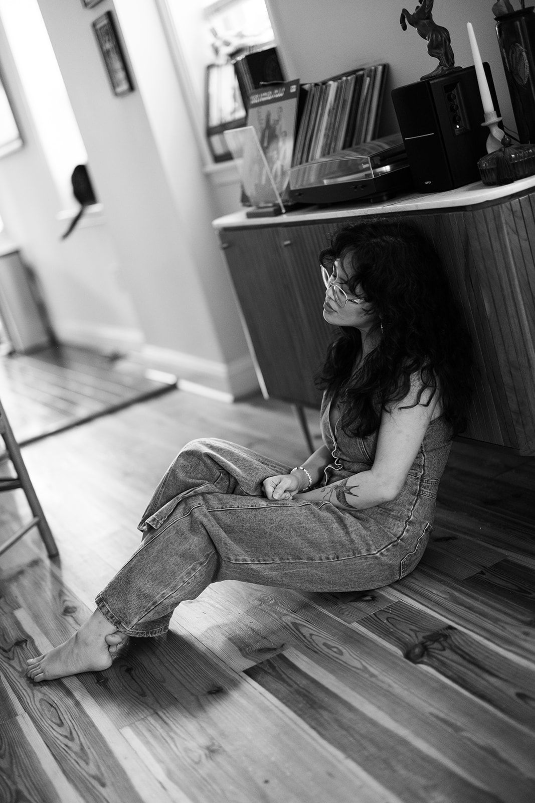 Black and white photo of a woman with curly hair and glasses sitting barefoot on a wood floor beside a console with records and a turntable.