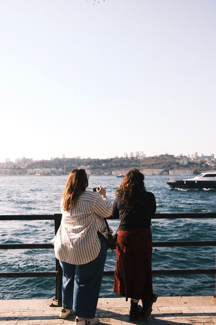 Two women stand at a waterfront railing photographing a scenic bay with boats and city skyline in the background.
