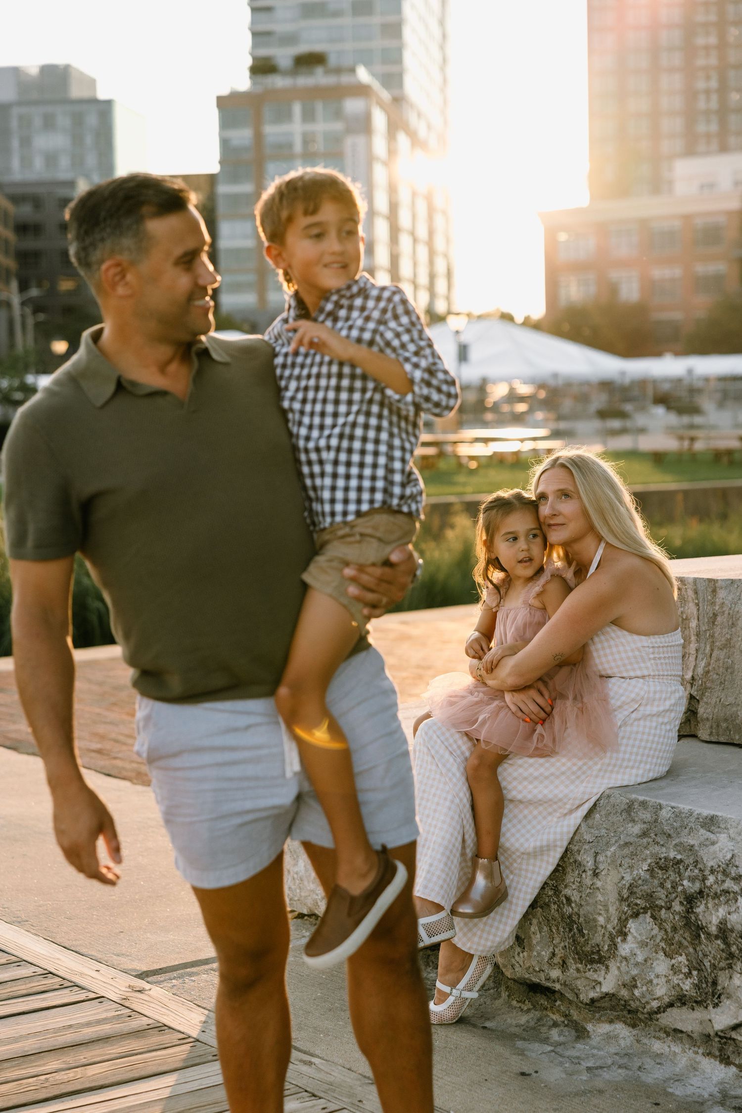 A happy family of four enjoys a golden sunset in an urban park, with dad holding son and mom hugging daughter.