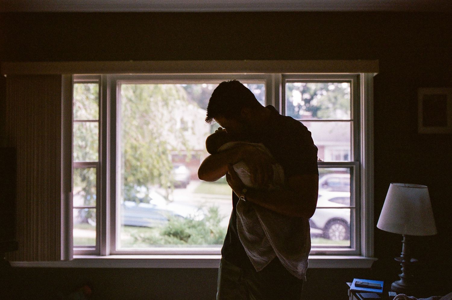 Silhouette of person cradling a baby by a bright window in a dimly lit room with a lamp nearby.