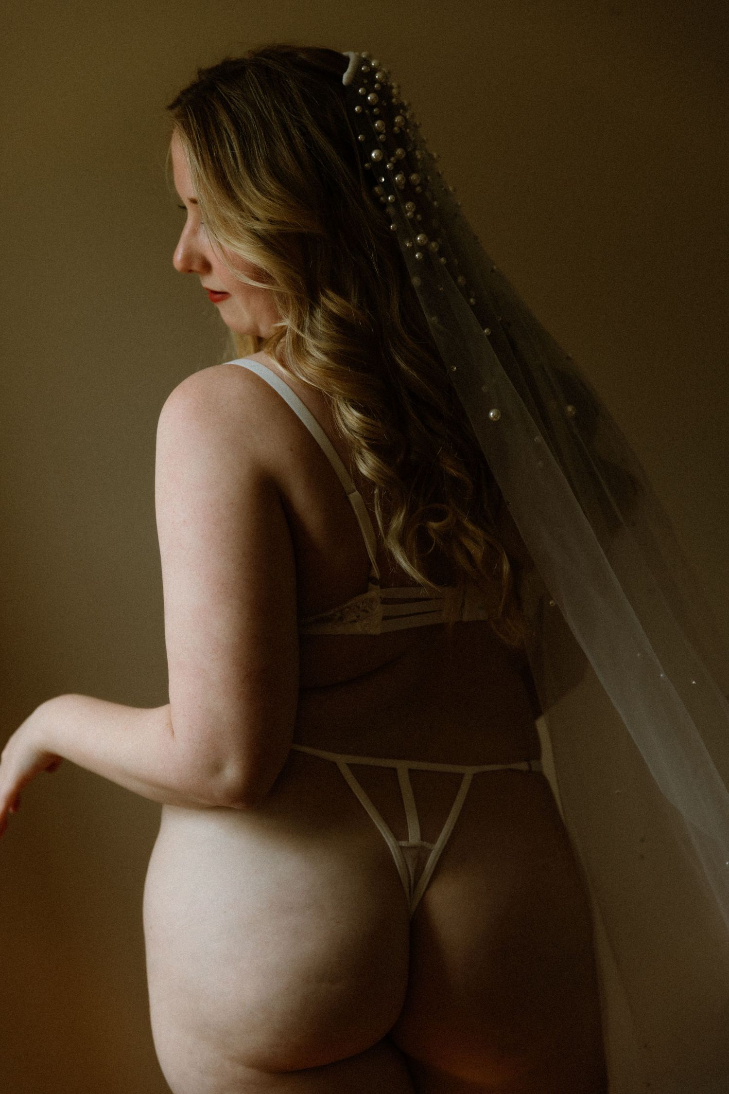 Bride in sheer lingerie and pearl-adorned veil poses elegantly in moody boudoir portrait photography.