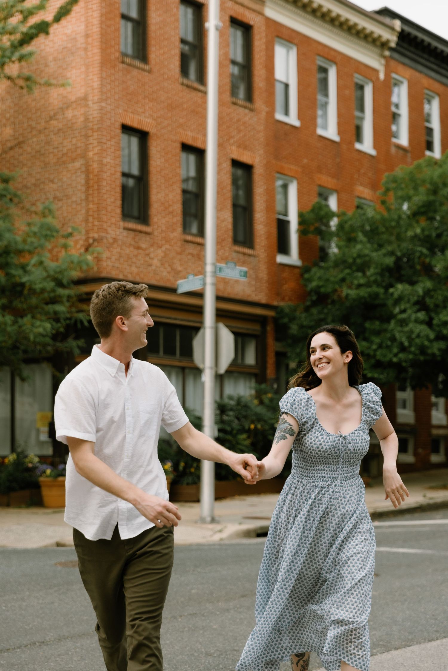 A smiling couple holds hands while walking through a charming urban street with brick buildings and lush trees.