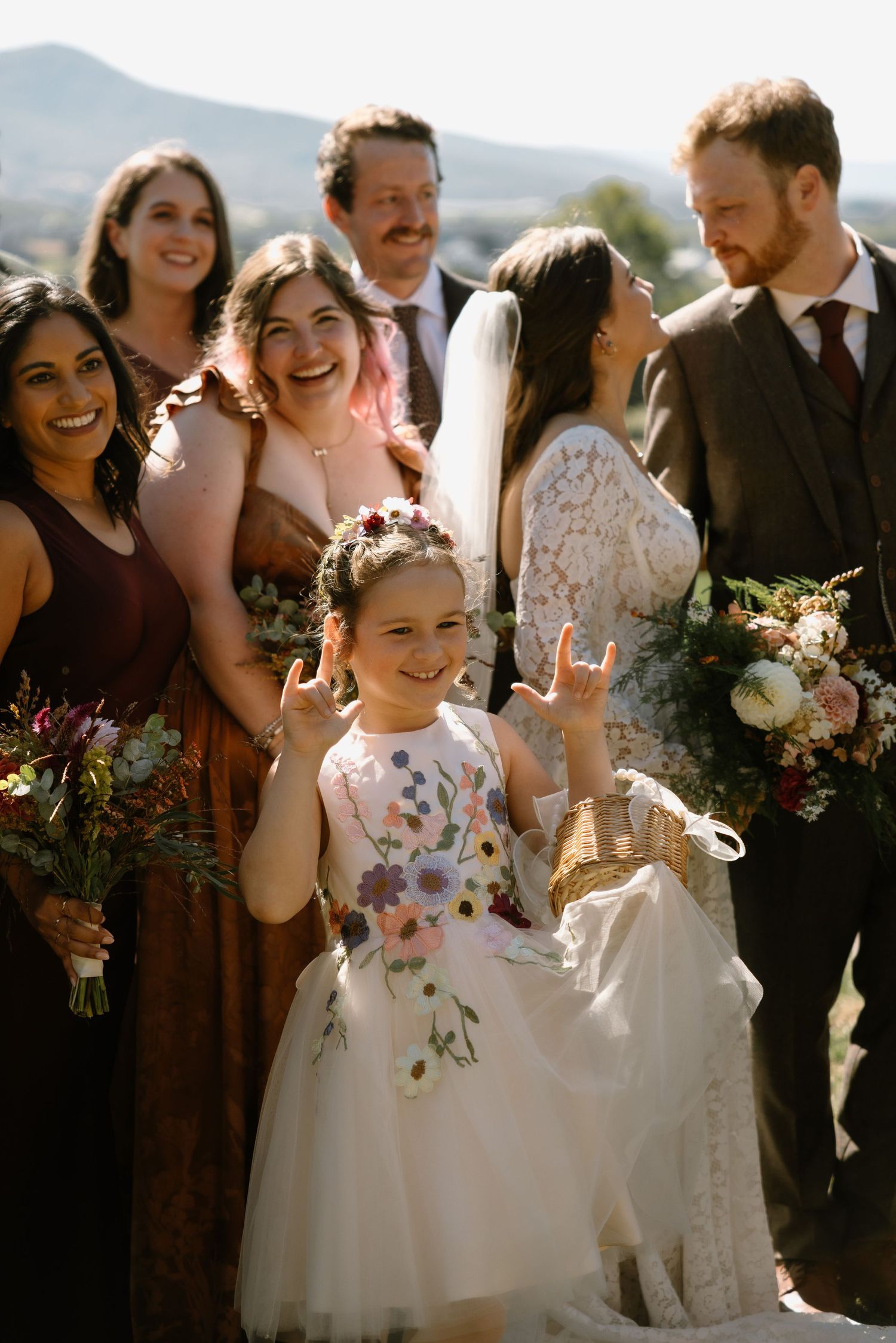 A flower girl flashes rock on signs at a wedding, surrounded by bridesmaids, groomsmen, bride and groom outdoors.