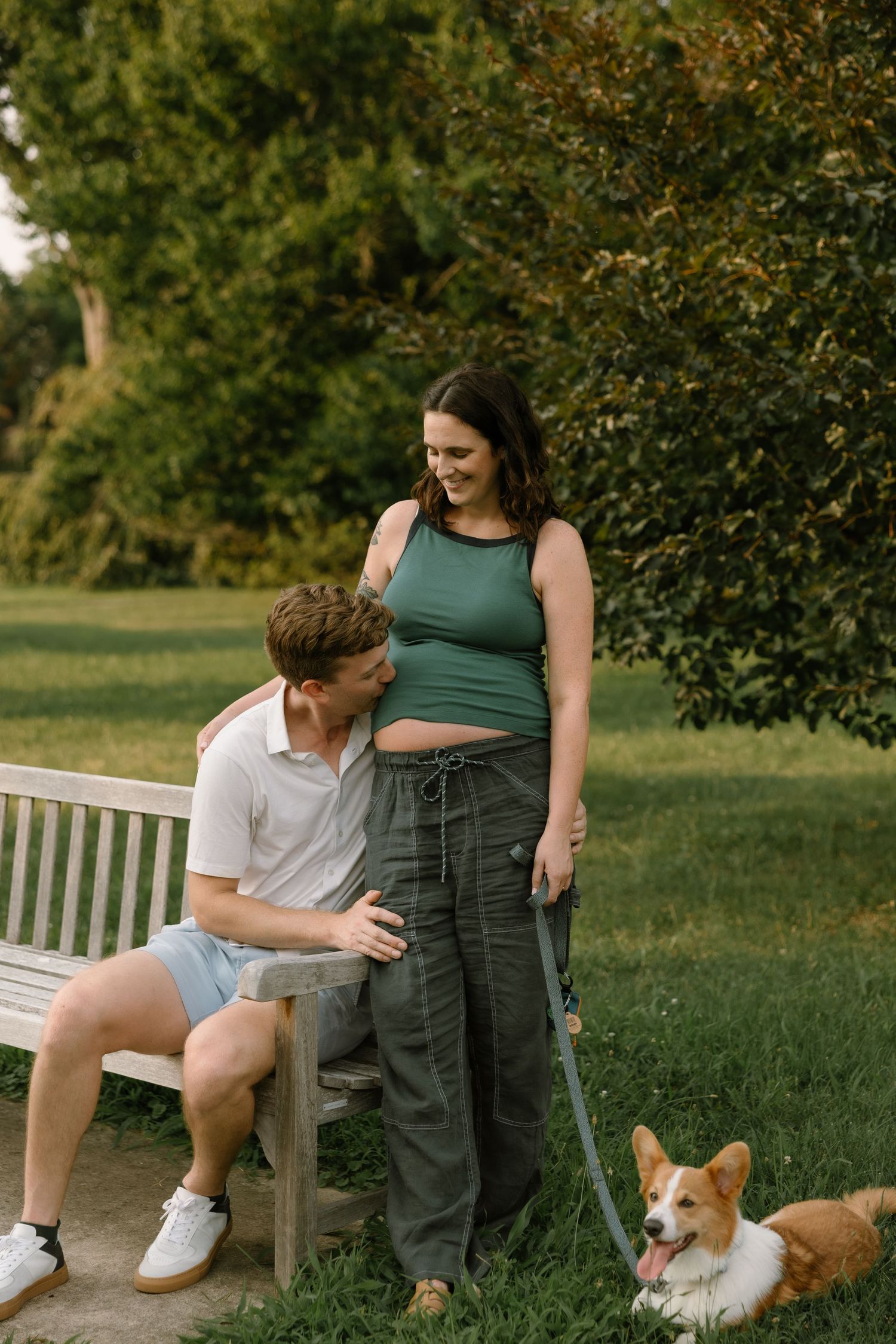 A couple with their Corgi dog relaxing on a park bench surrounded by lush green trees on a sunny day.