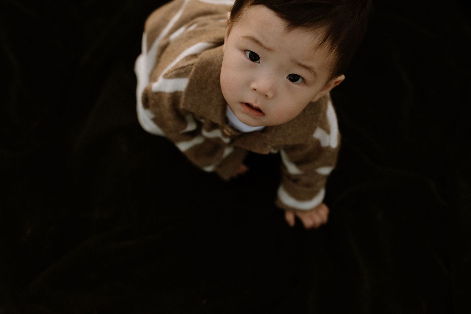 A cute toddler in a brown patterned sweater looks up at the camera from a dark floor.