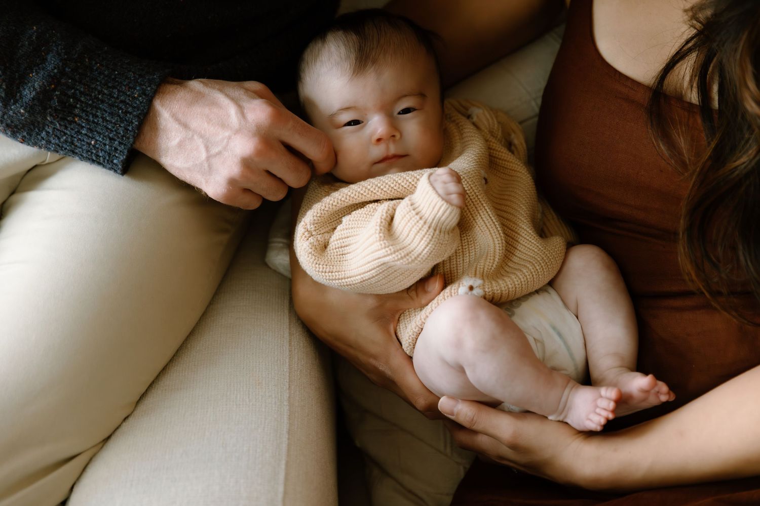 Newborn baby in beige knit outfit resting between parents on a cream sofa during a family lifestyle photo session.