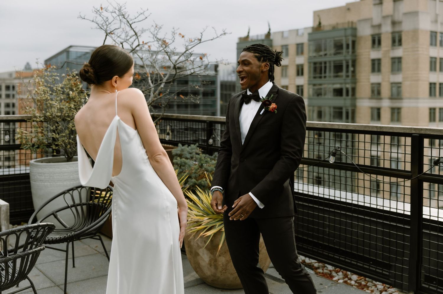 Bride in elegant backless white gown and groom in black tuxedo share a joyful moment on a rooftop terrace.