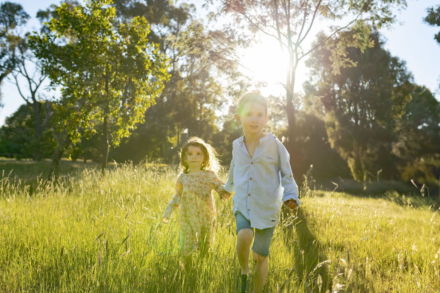 Brother and sister holding hands walking in a field of tall grass with warm sunlight looking at each other, outdoor home Woodend , Melb, Vic