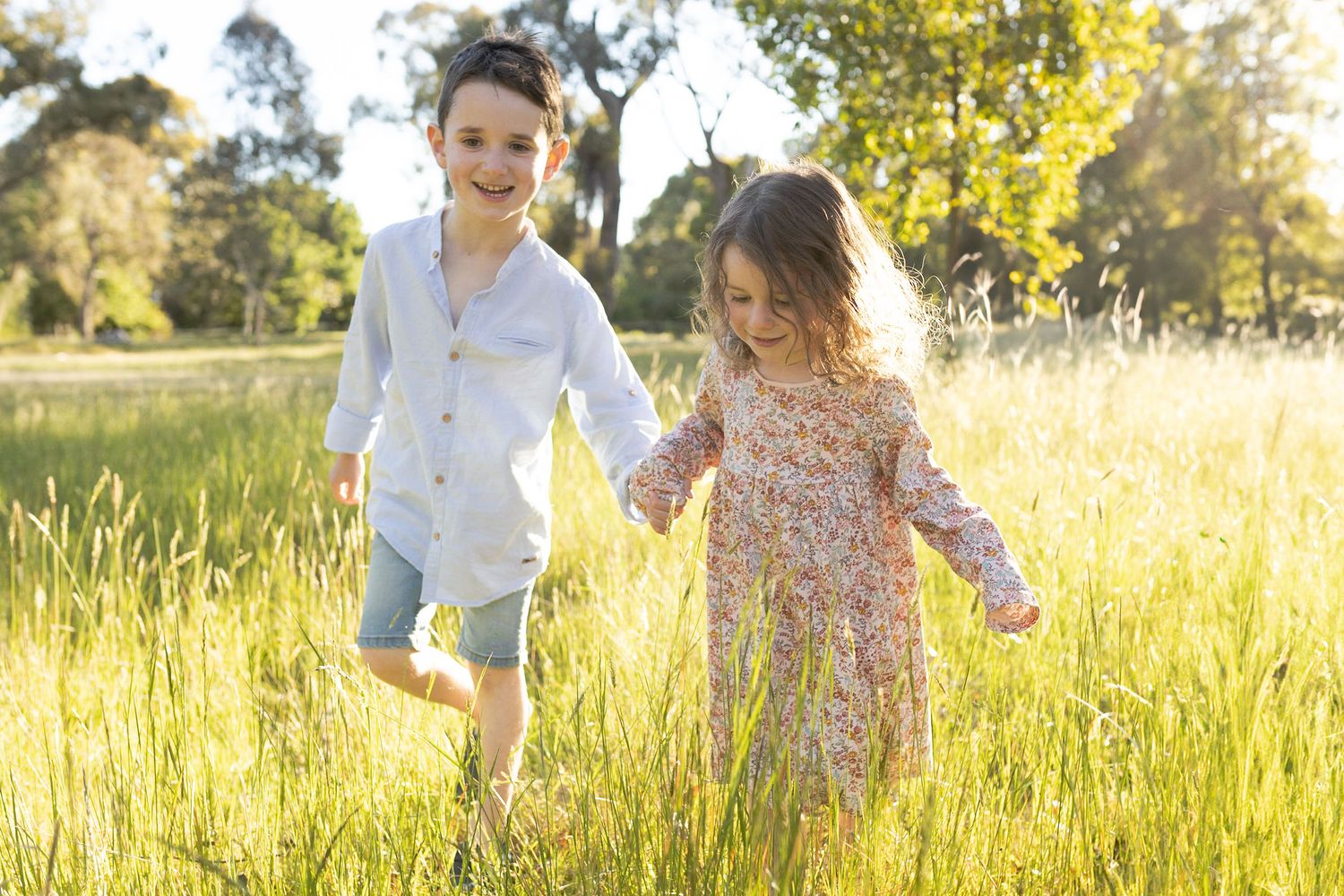 Brother and sister holding hands walking in a field of tall grass with warm sunlight looking at each other, outdoor home Woodend , Melb, Vic