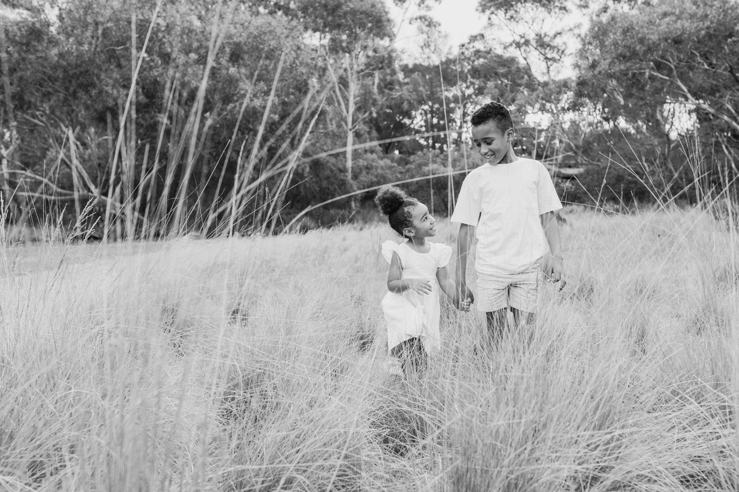 Brother and sister holding hands walking in a field of tall grass with warm sunlight looking at each other, Newport Lakes, Melb, Vic
