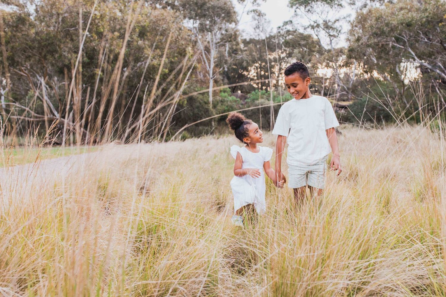 Brother and sister holding hands walking in a field of tall grass with warm sunlight looking at each other, Newport Lakes, Melb, Vic