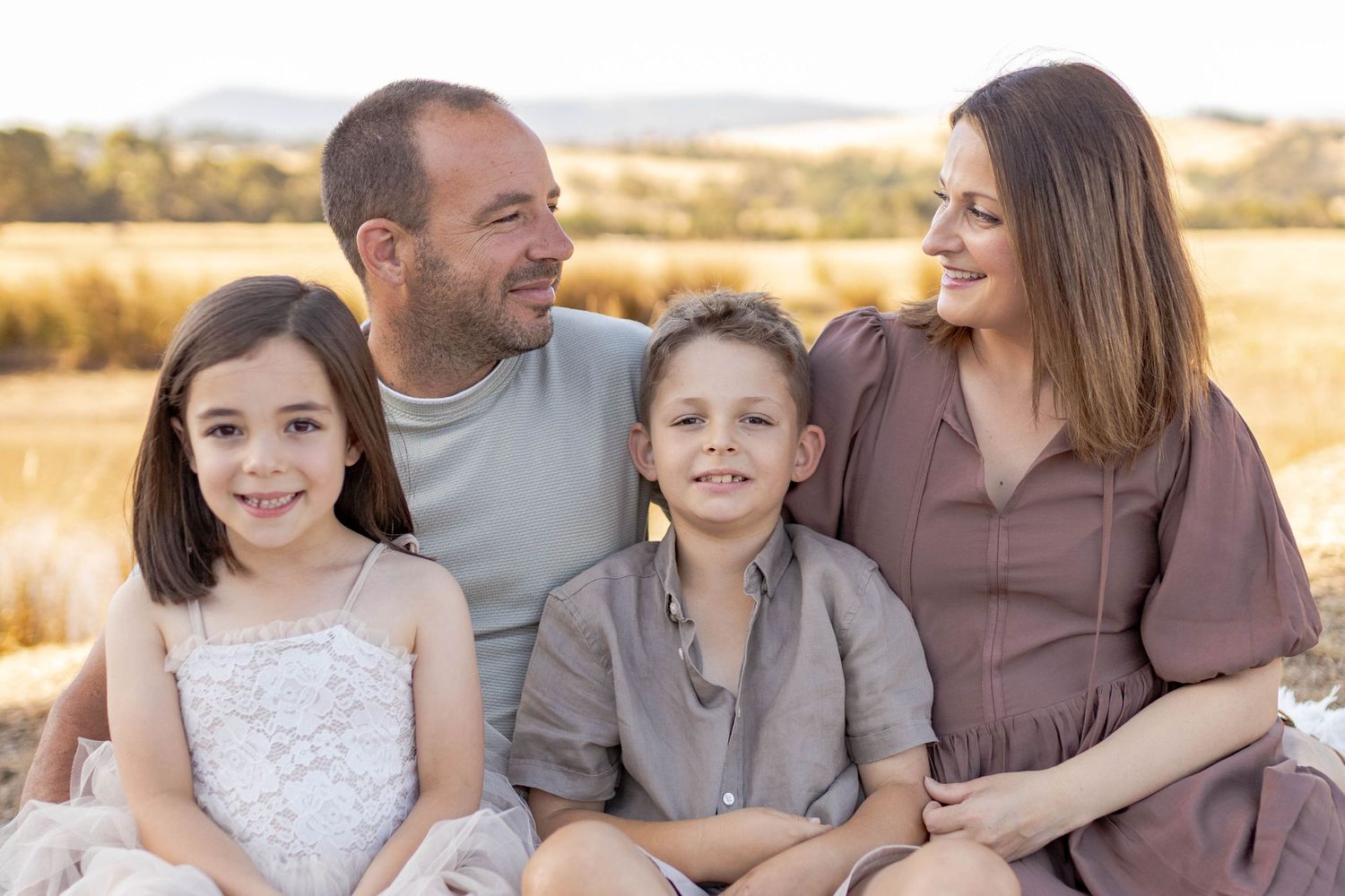Family of four sitting on a white blanket on a hill with fields in the background Outdoor at home family session Gisborne Melb Vic