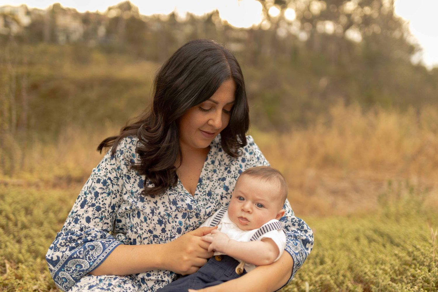 Mother holding baby boy in her arms and looking down with a grin warm light. Outdoor family session Sunshine North Melb Vic