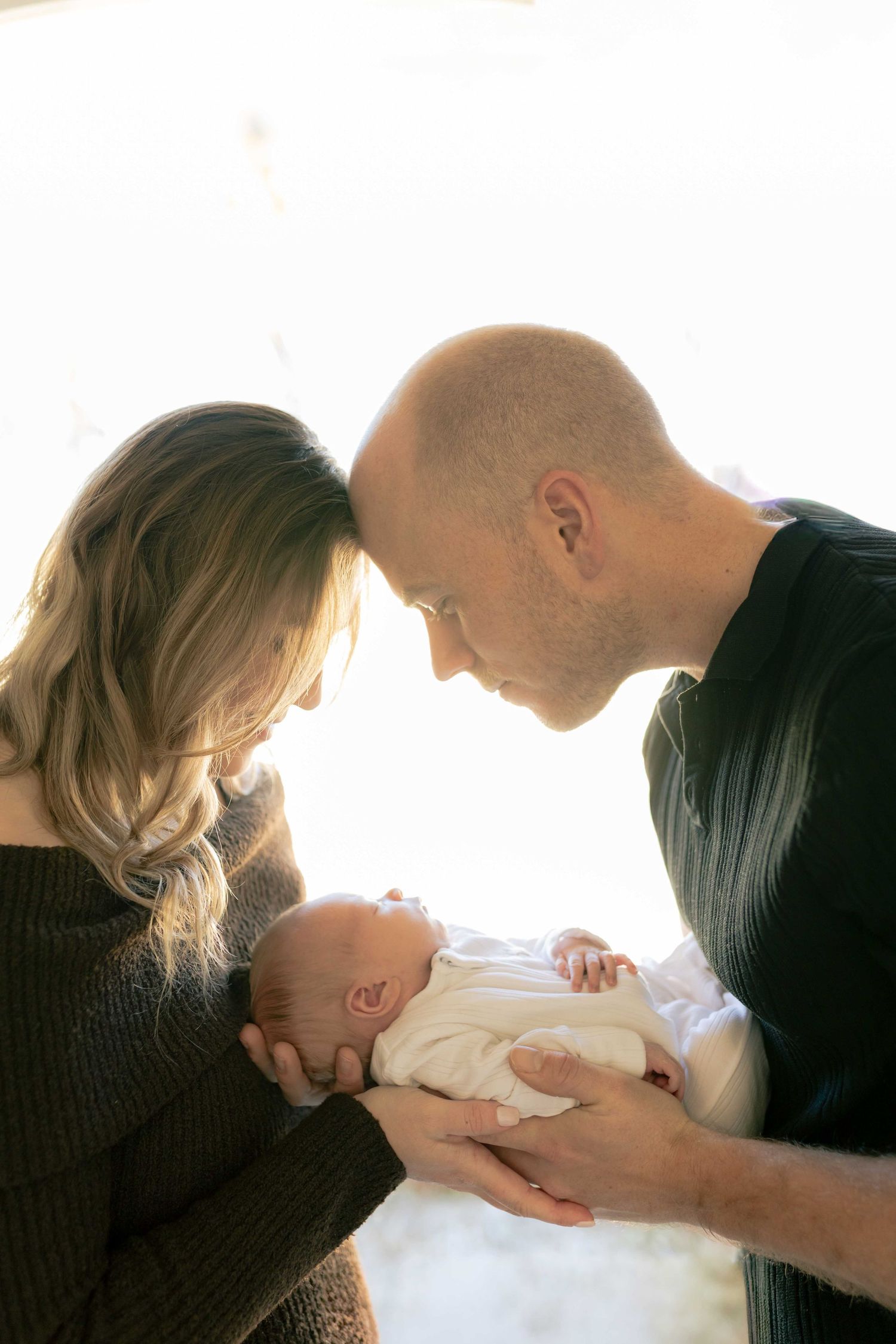 Mum and dad gently pressing their heads together looking down at their newborn baby between them. Newborn Photoshoot Melbourne's West Melton