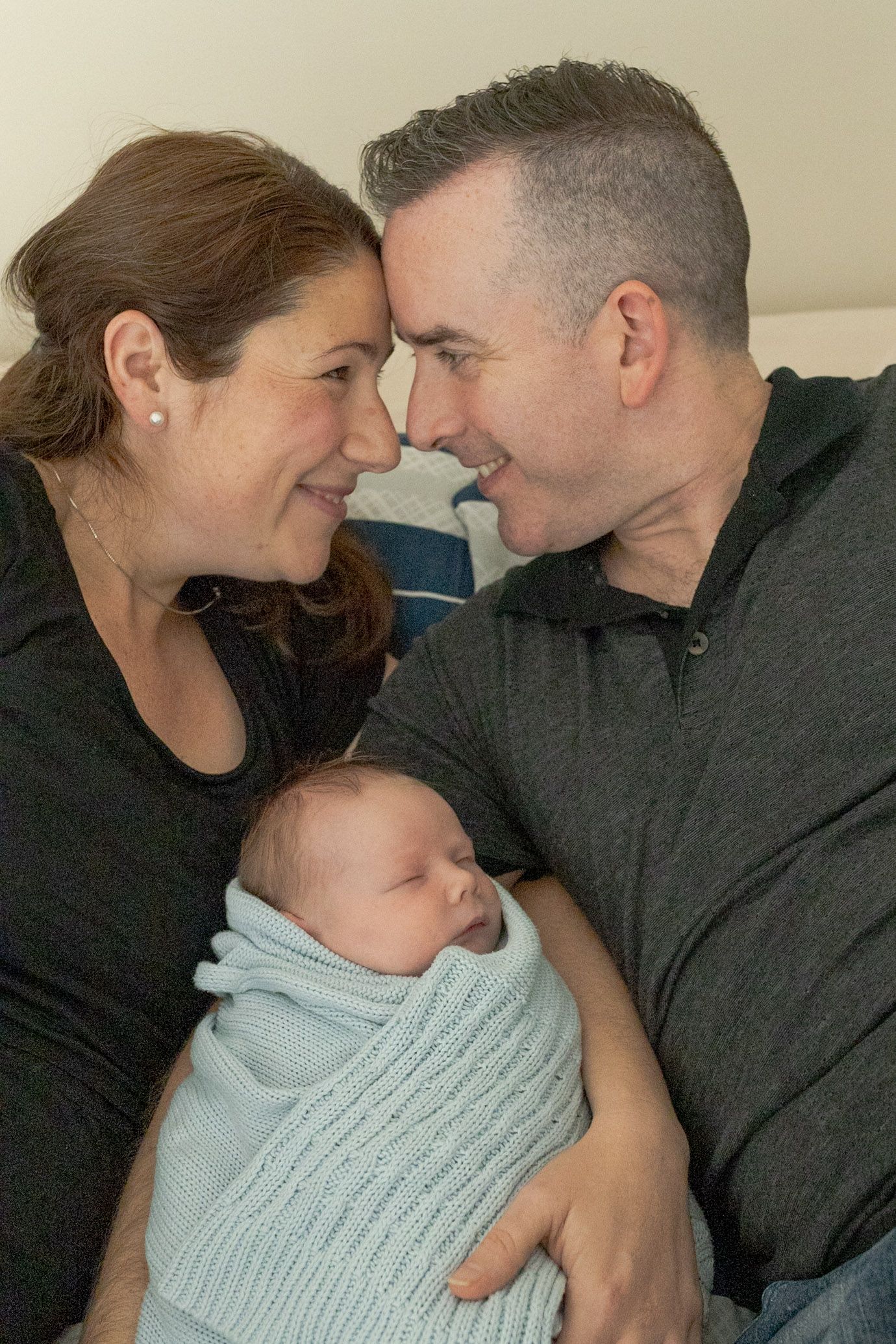 Parents snuggling newborn baby boy while lying on the bed baby swaddled in blue blanket at home newborn session  Moonee Ponds Melb Vic