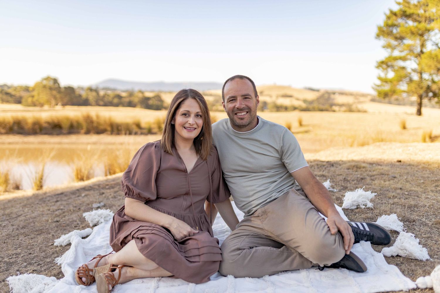 Mum and dad sitting on a white blanket on a hill with fields in the background Outdoor at home family session Gisborne Melb Vic