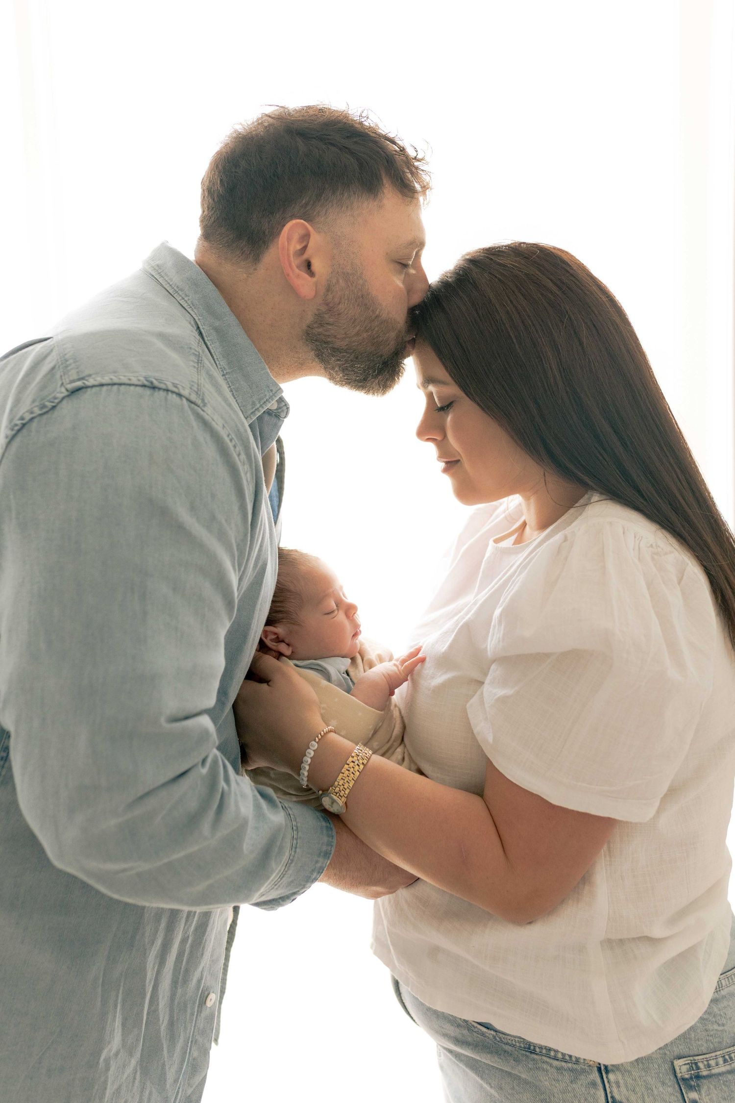 Dad reaching over and kissing mum on the head who is looking down at their baby
