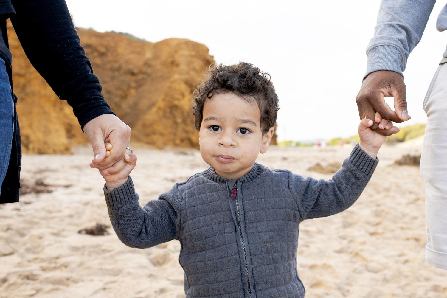 Toddler holding onto his parents hands and walking on the sand at the beach Outdoor family session Torquay