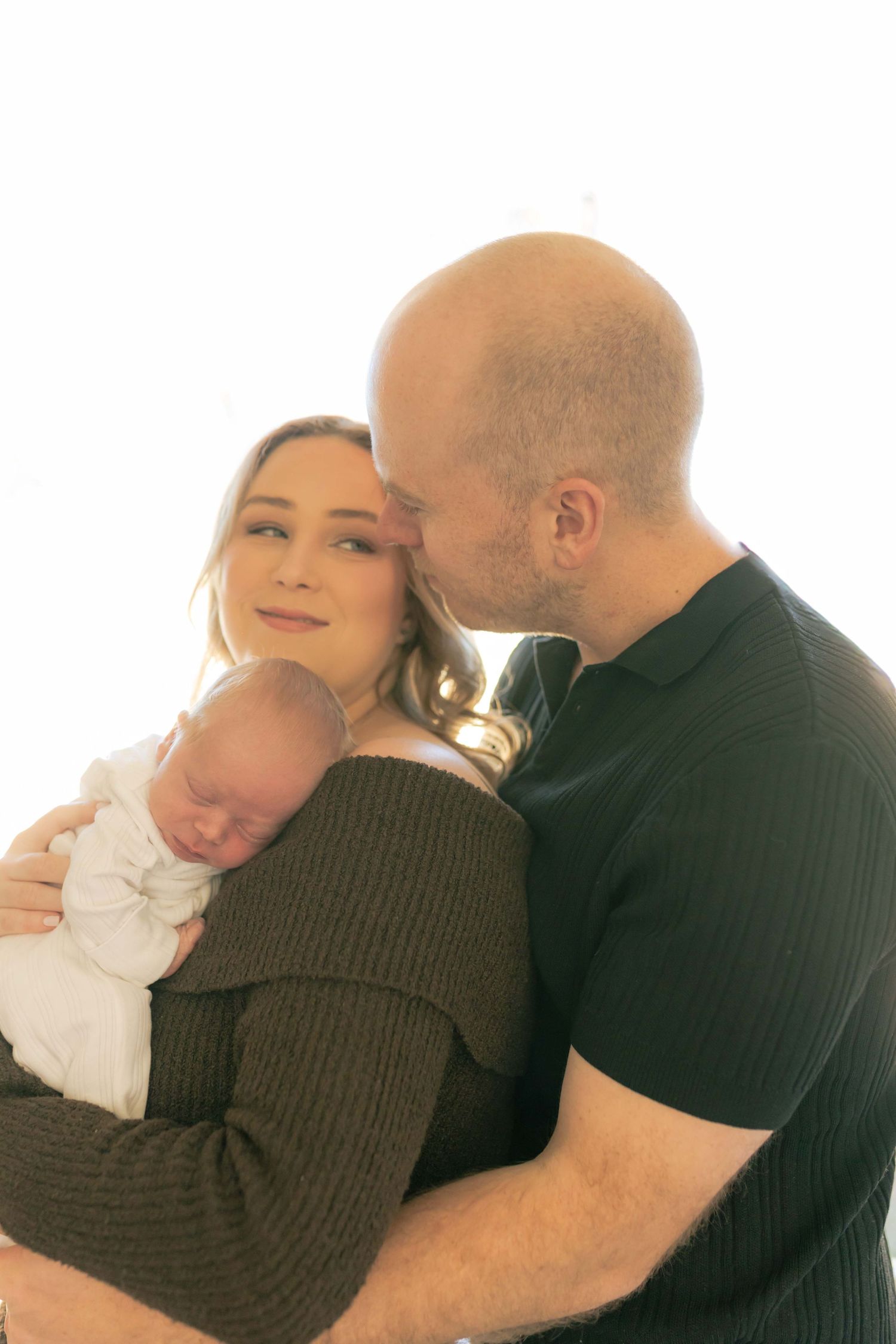Mum holding newborn boy with dad hugging mum from behind