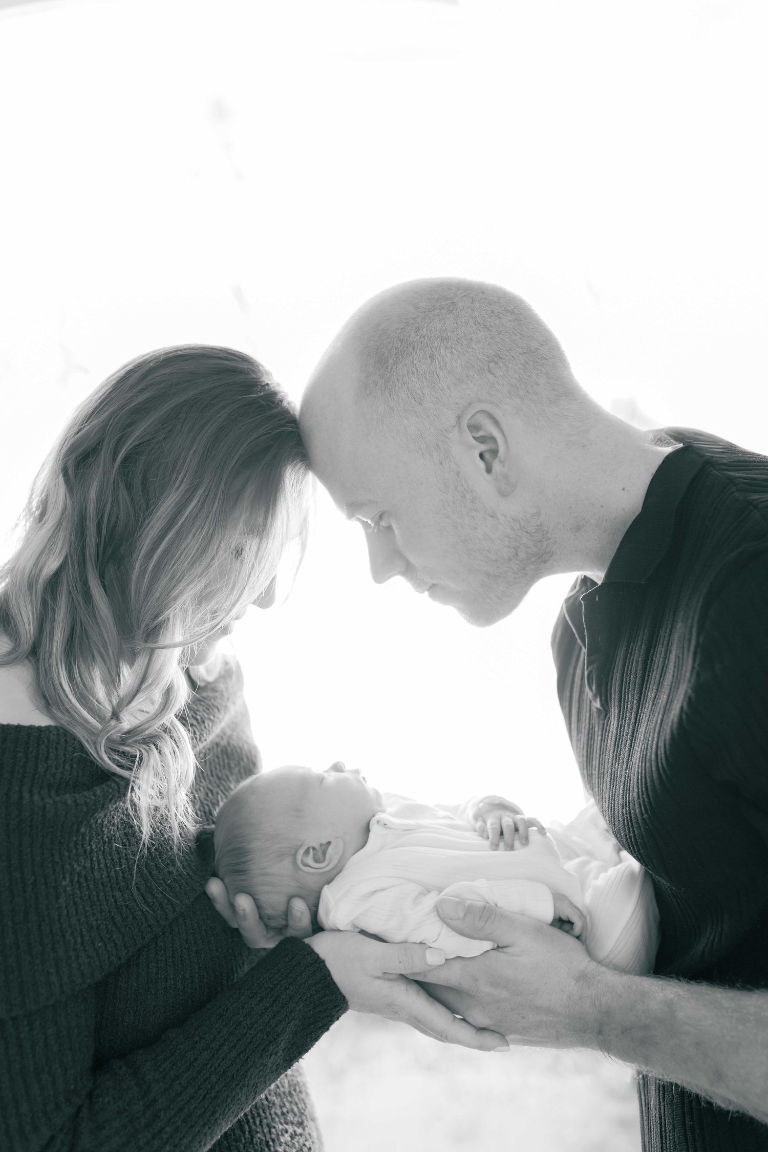 Mum and dad gently pressing their heads together looking down at their newborn baby between them. Newborn Photoshoot Melbourne's West Melton