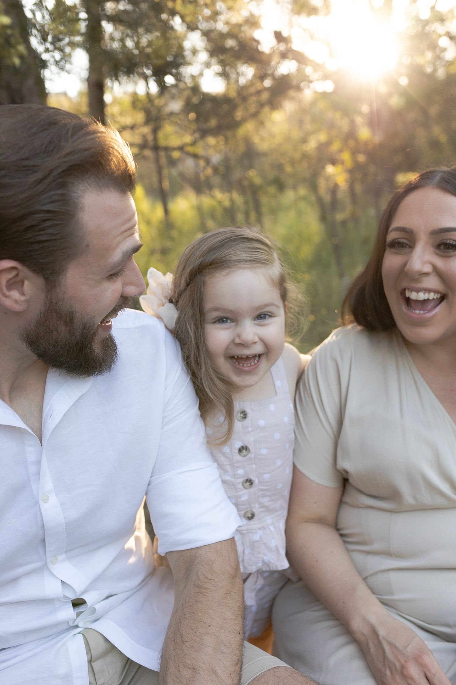 Little girl playing peek a boo from behind her mum and dad, smiling at each other outside in bushland, outdoor Sunshine North, Melb, Vic