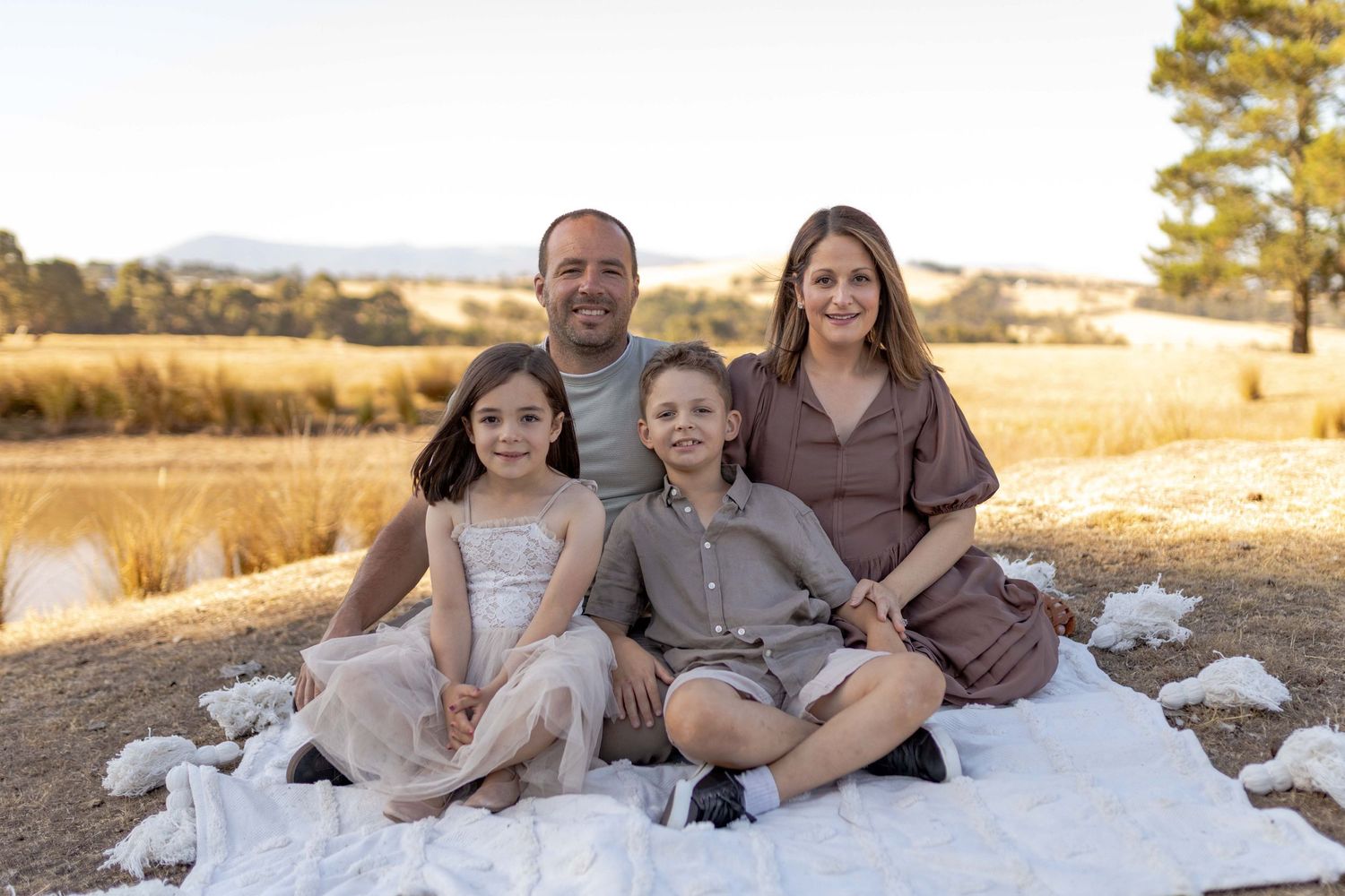 Family of four sitting on a white blanket on a hill with fields in the background Outdoor at home family session Gisborne Melb Vic