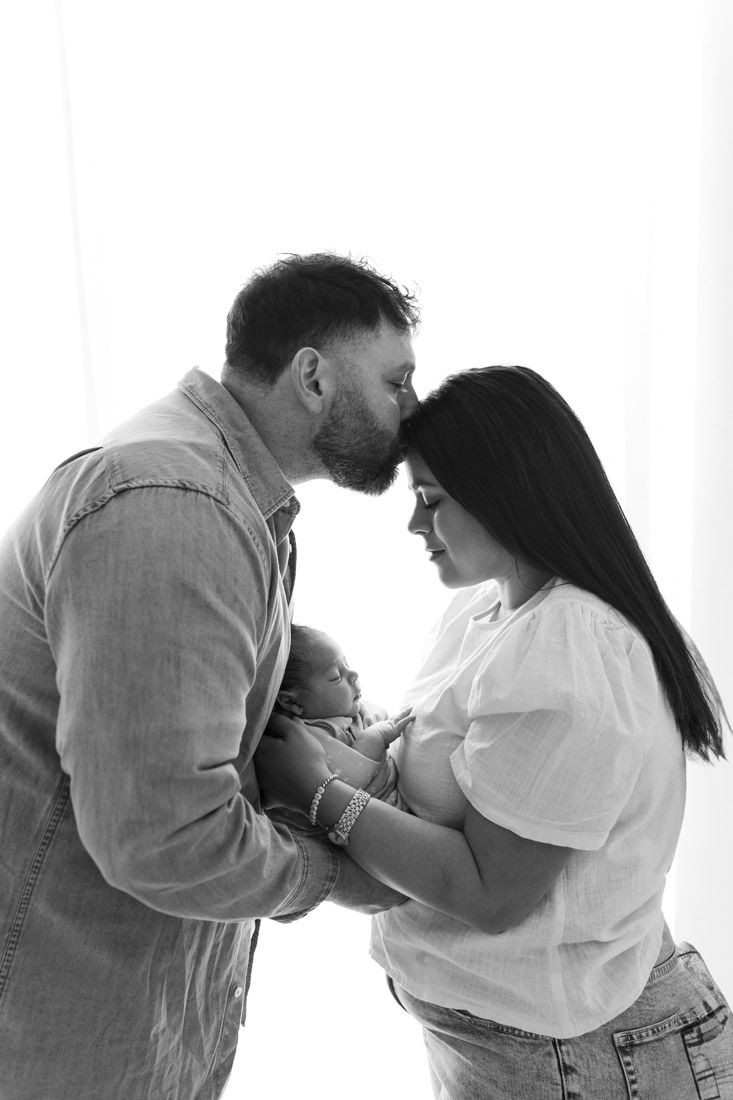 Dad reaching over and kissing mum on the head who is looking down at their baby