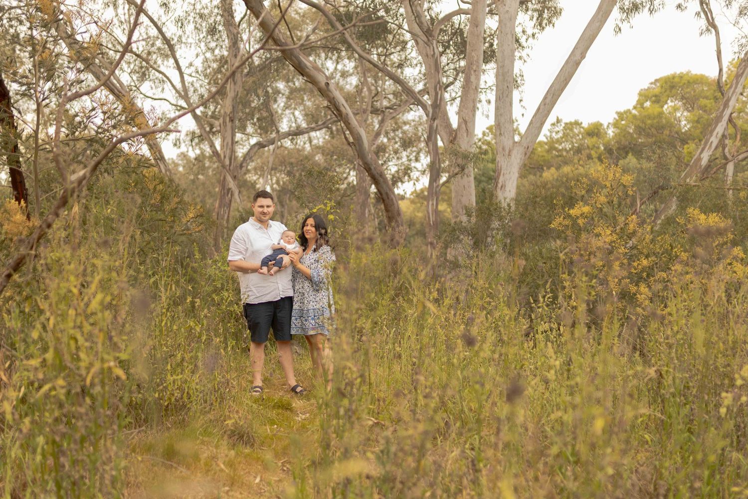 First time parents holding newborn in the bushland with tall grass along the path. Outdoor family session Sunshine North Melb Vic
