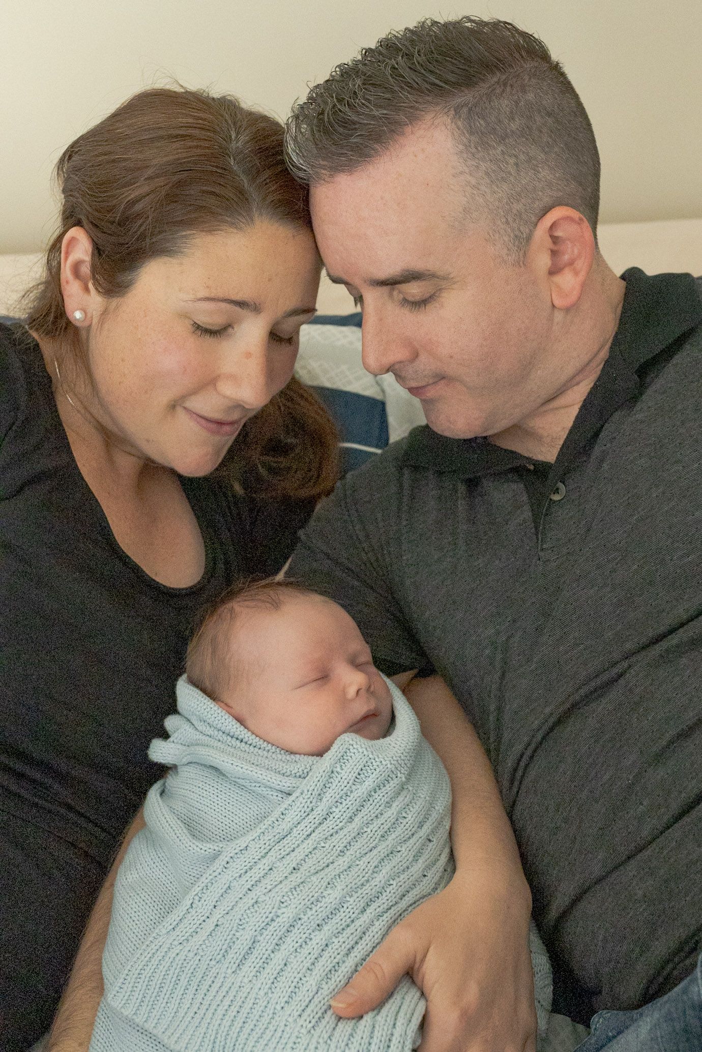 Parents snuggling newborn baby boy while lying on the bed baby swaddled in blue blanket at home newborn session  Moonee Ponds Melb Vic