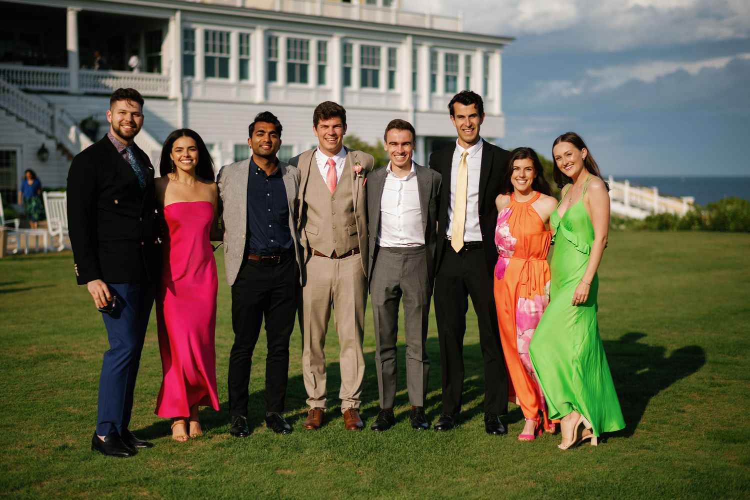A group of formally dressed people pose together on a green lawn with a white building and ocean view in the background.