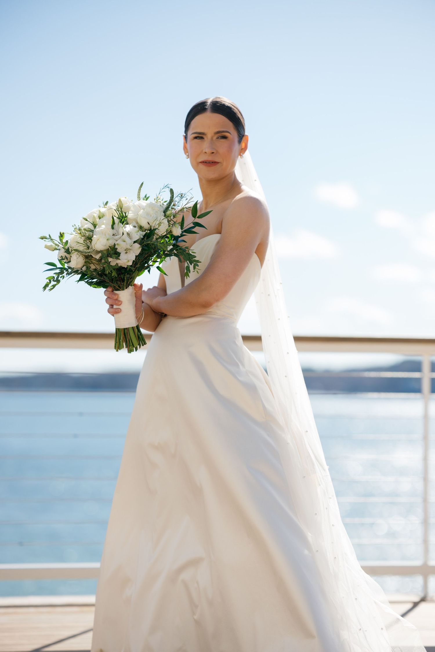 Elegant bride in white gown holding bouquet of white flowers, standing on waterfront deck with blue sky backdrop.