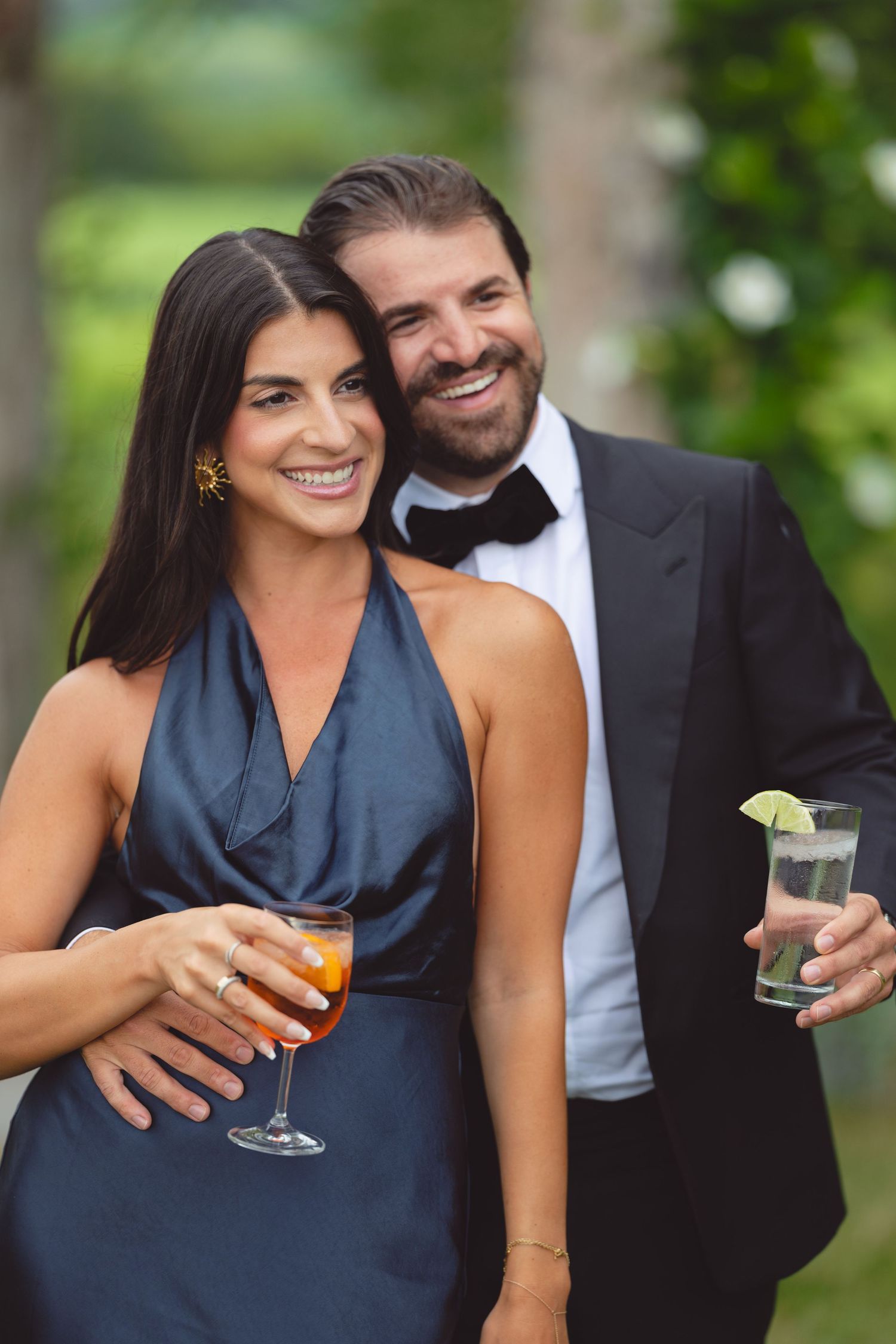 Couple laughing and sharing drinks at outdoor celebration in elegant navy dress and dark suit.