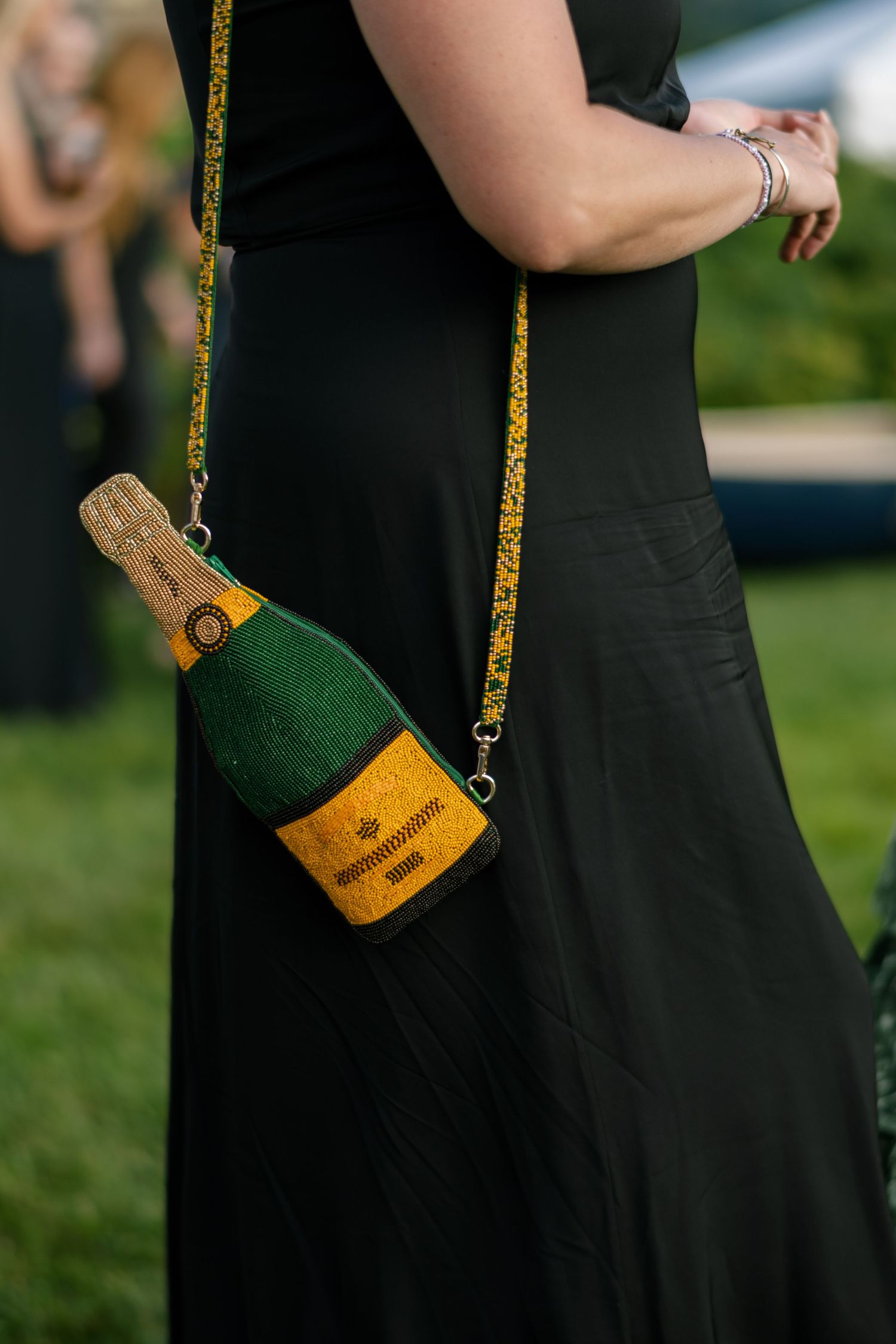 A novelty champagne bottle purse with gold strap hangs against a black dress at an outdoor event.