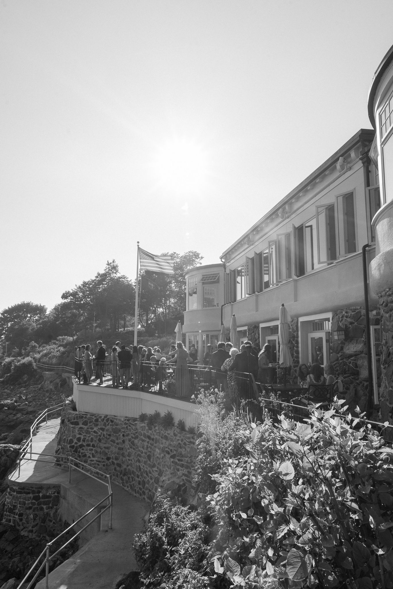 Wedding guests gather outside a building under bright sunlight with an American flag visible.