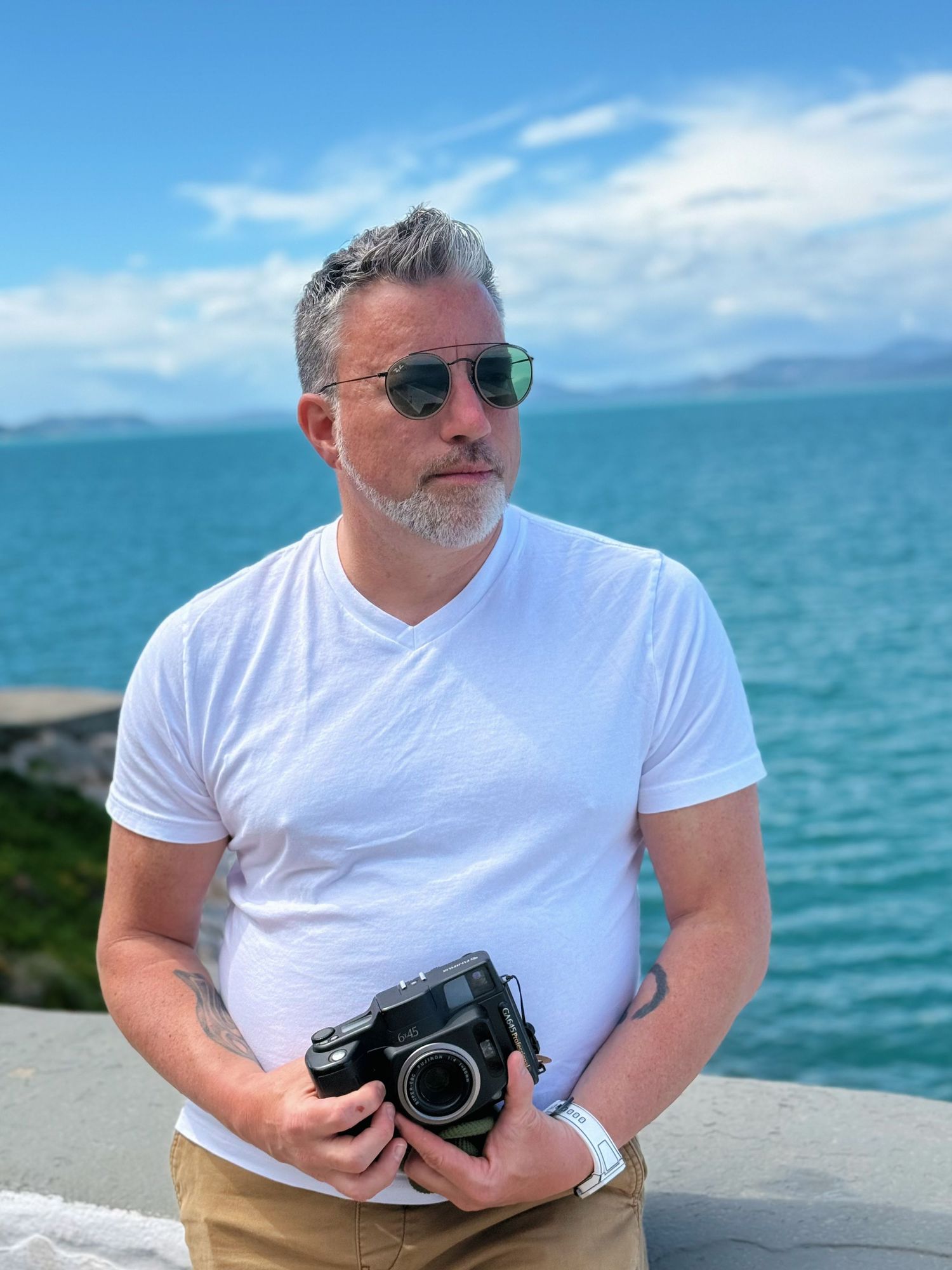 Man in white t-shirt holding vintage camera by turquoise ocean, wearing sunglasses on sunny day.