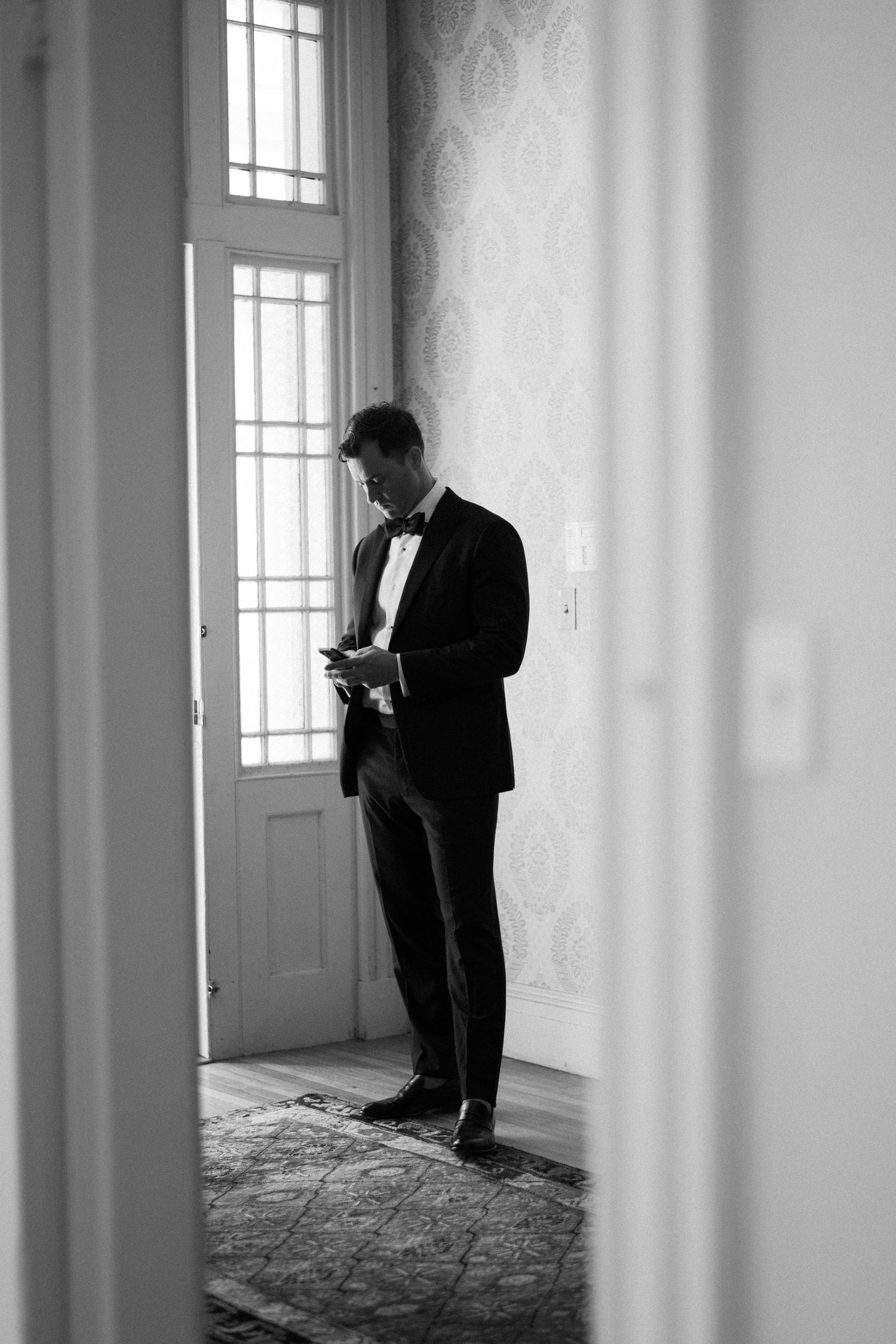 Black and white photo of a groom in a tuxedo and bow tie checking his phone in a elegant room before the wedding ceremony.